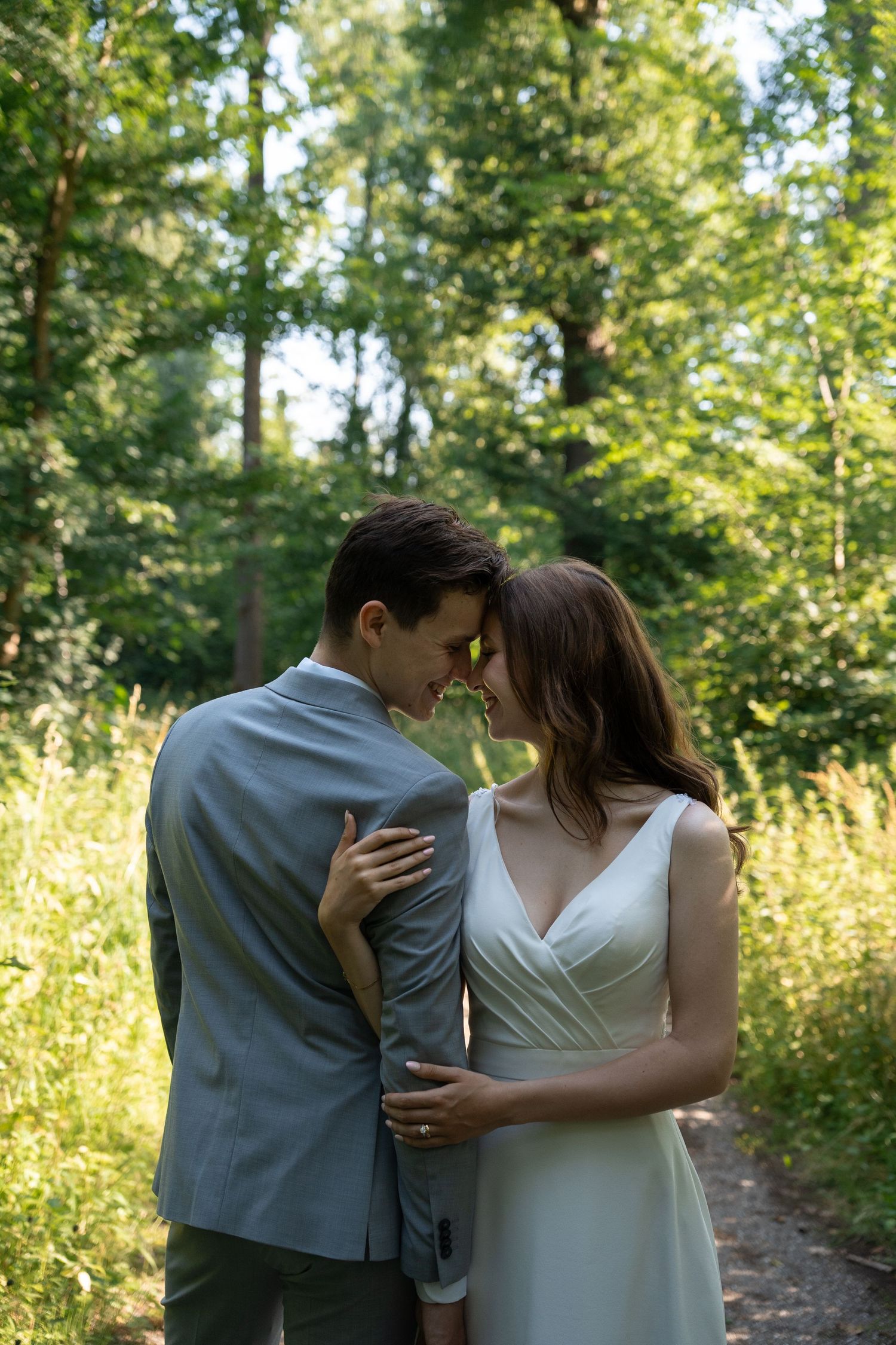 Bride and groom sharing intimate moment on forest path in soft natural light.