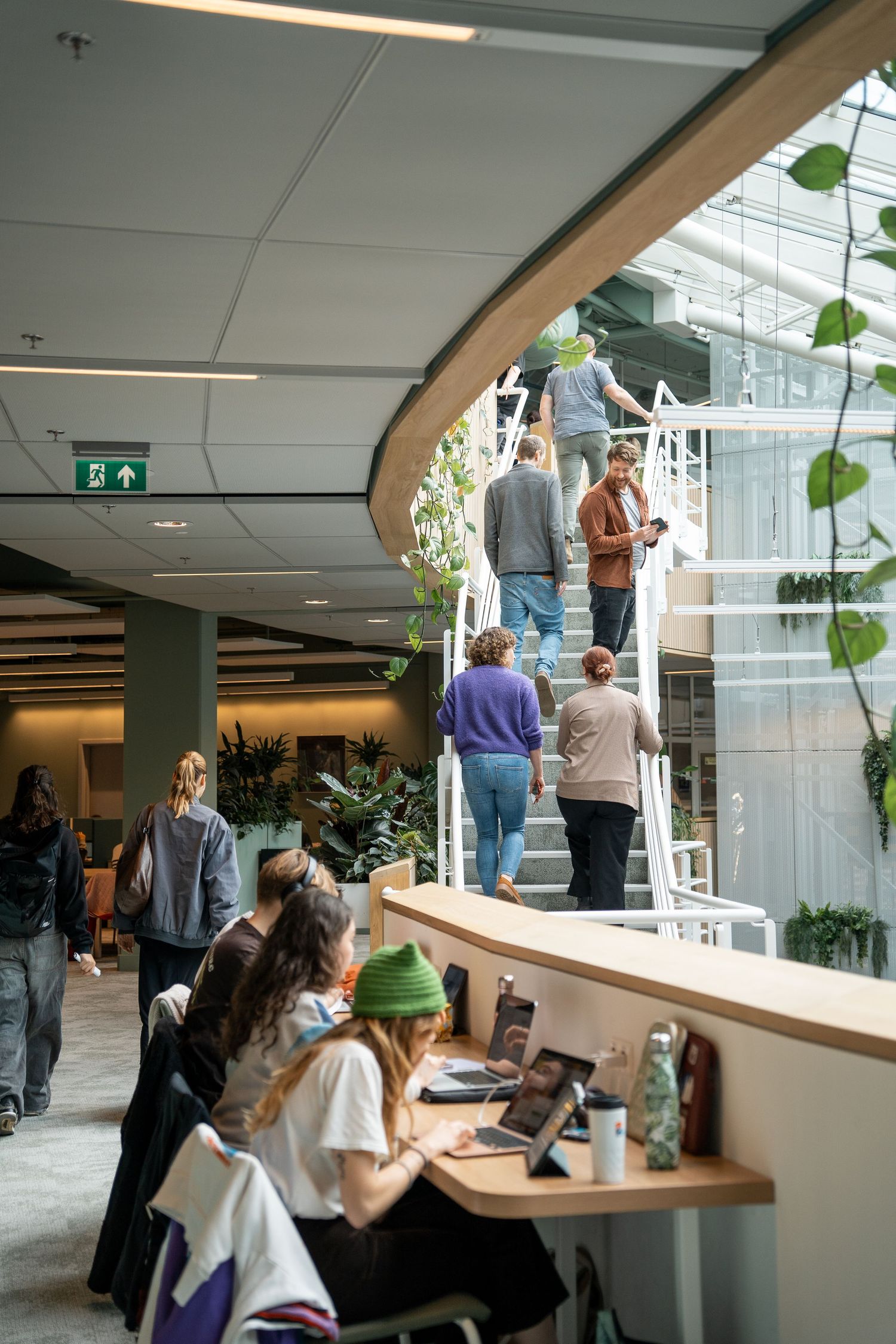 Students studying and walking in modern university building with plants and spiral staircase.