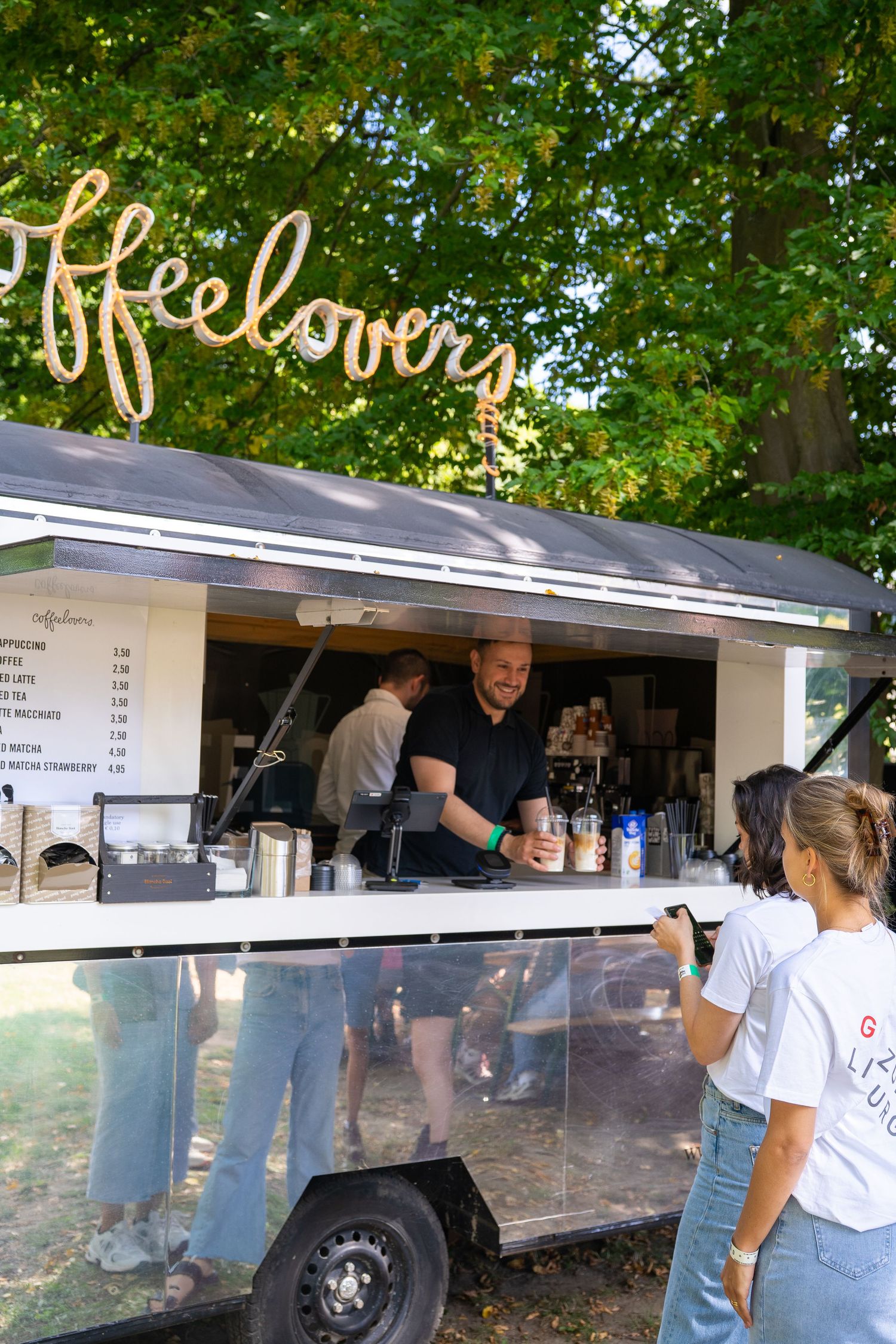 A barista serves coffee at an outdoor coffeelovers food truck booth surrounded by trees.