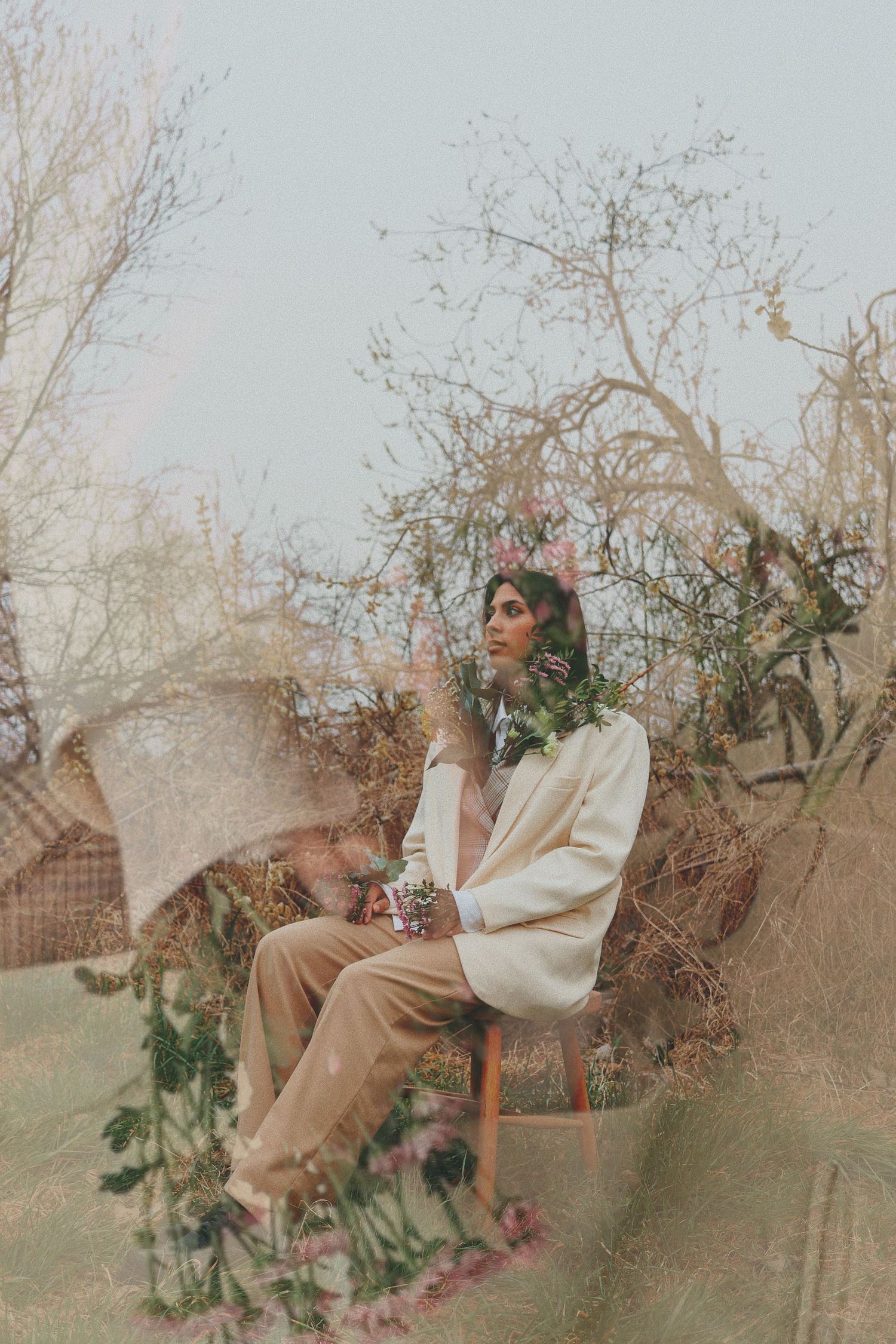 Woman in cream blazer sits on chair surrounded by wildflowers and bare trees in a dreamy double exposure outdoor portrait.