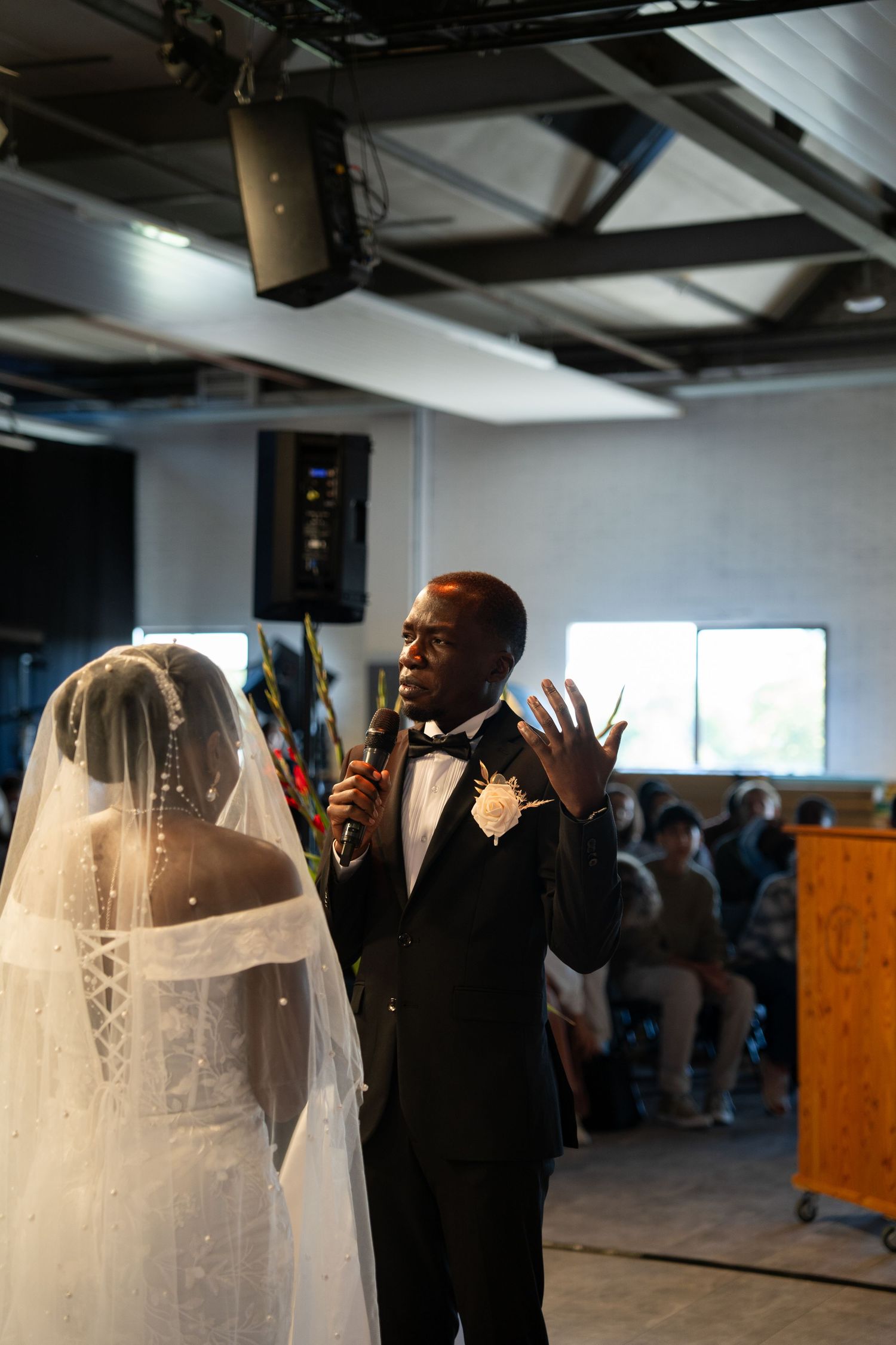 Groom in black tuxedo speaking vows into microphone facing bride in white gown during wedding ceremony.