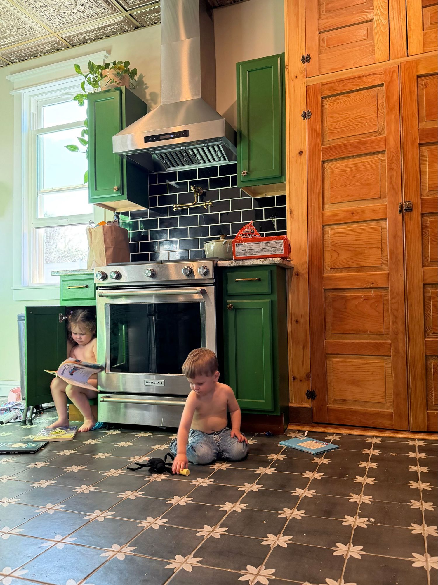 Two young children playing on a decorative tile floor in a kitchen with green cabinets and stainless steel range.