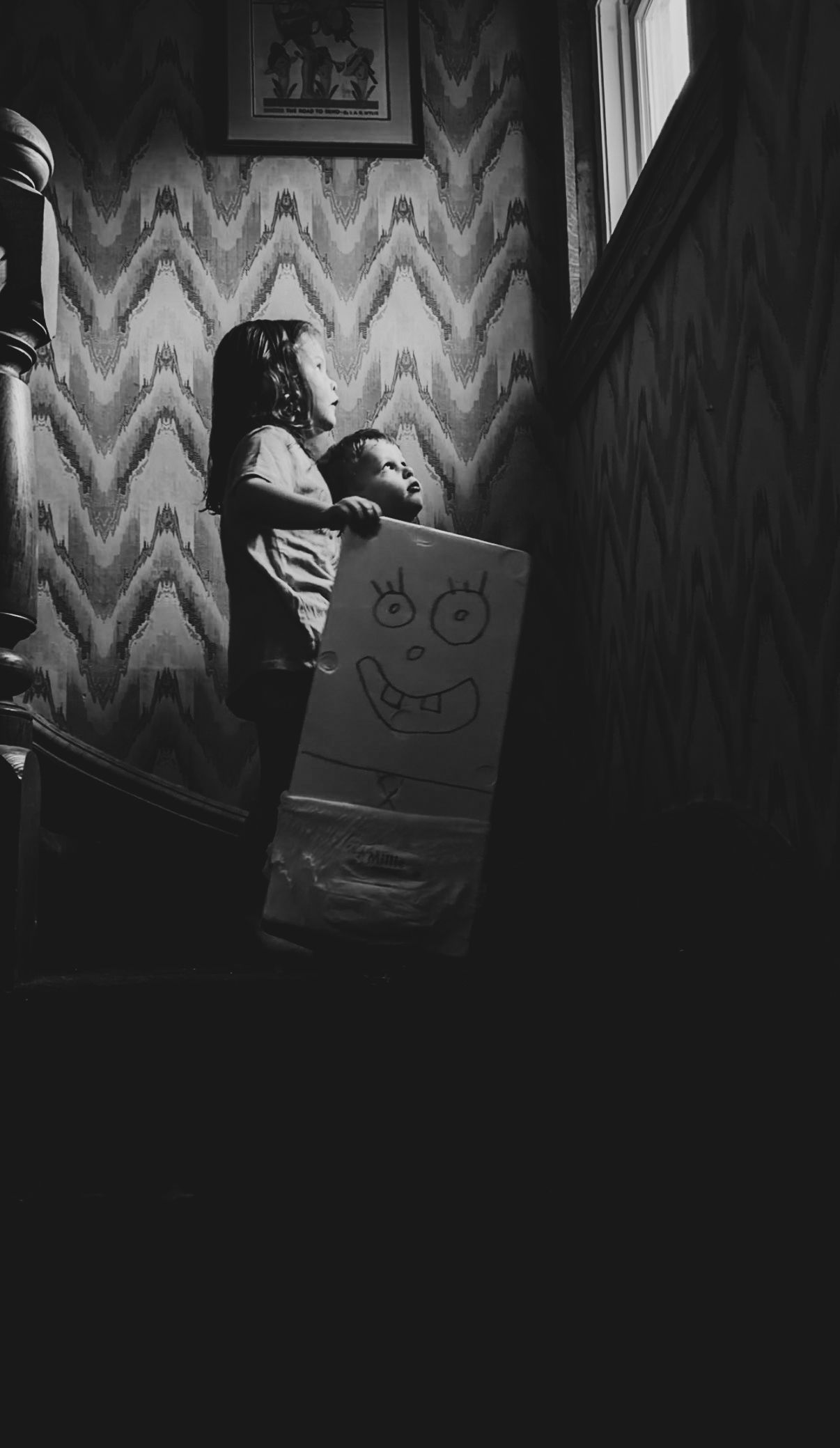 Black and white photo of a child holding a cardboard box with a monster face drawing on a staircase.