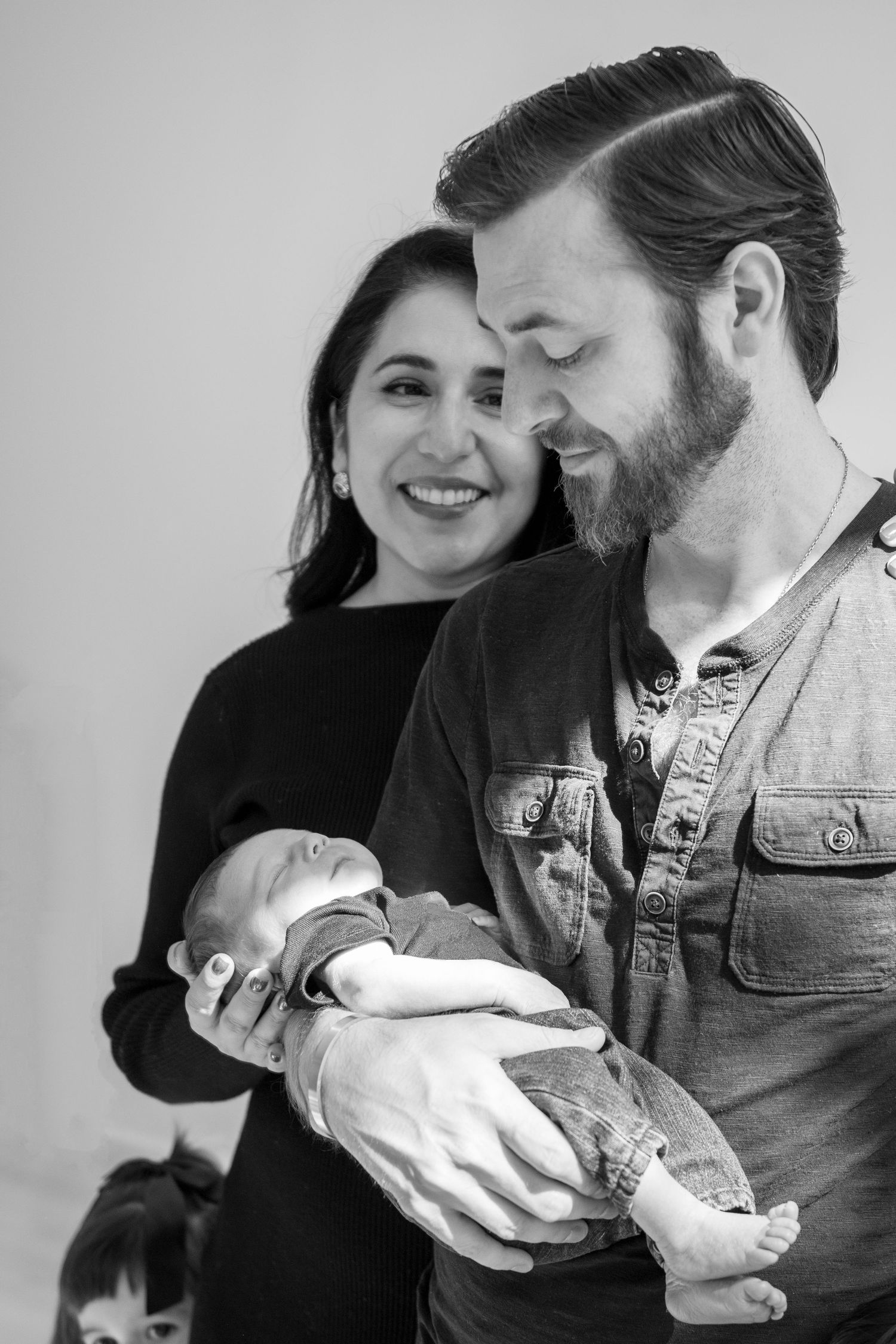 Smiling couple with bearded man holding newborn baby in black and white family portrait.