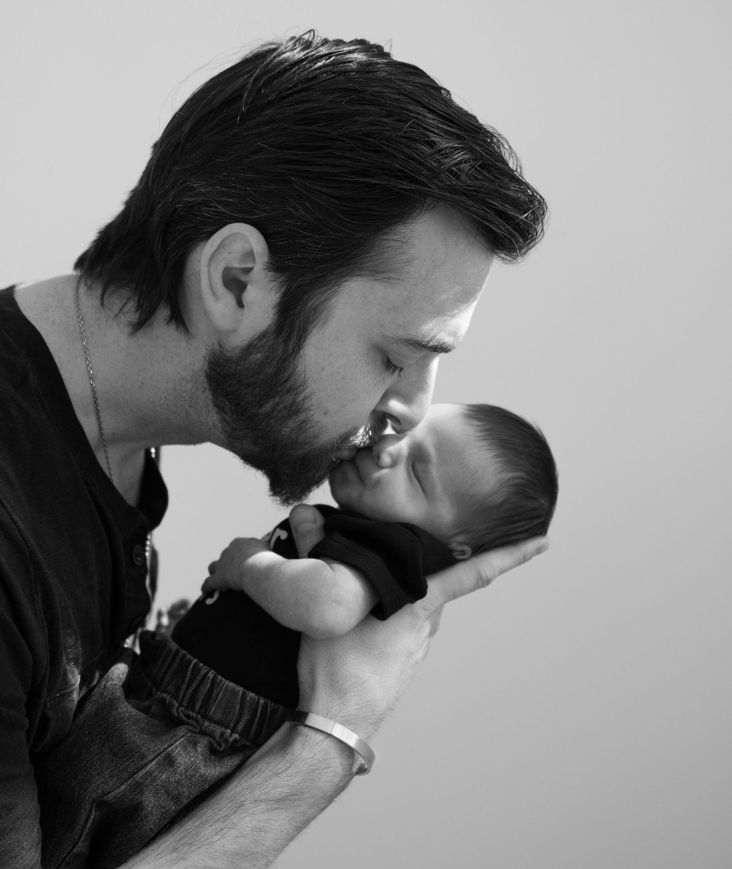 Tender black and white portrait of bearded father gently kissing his newborn baby.