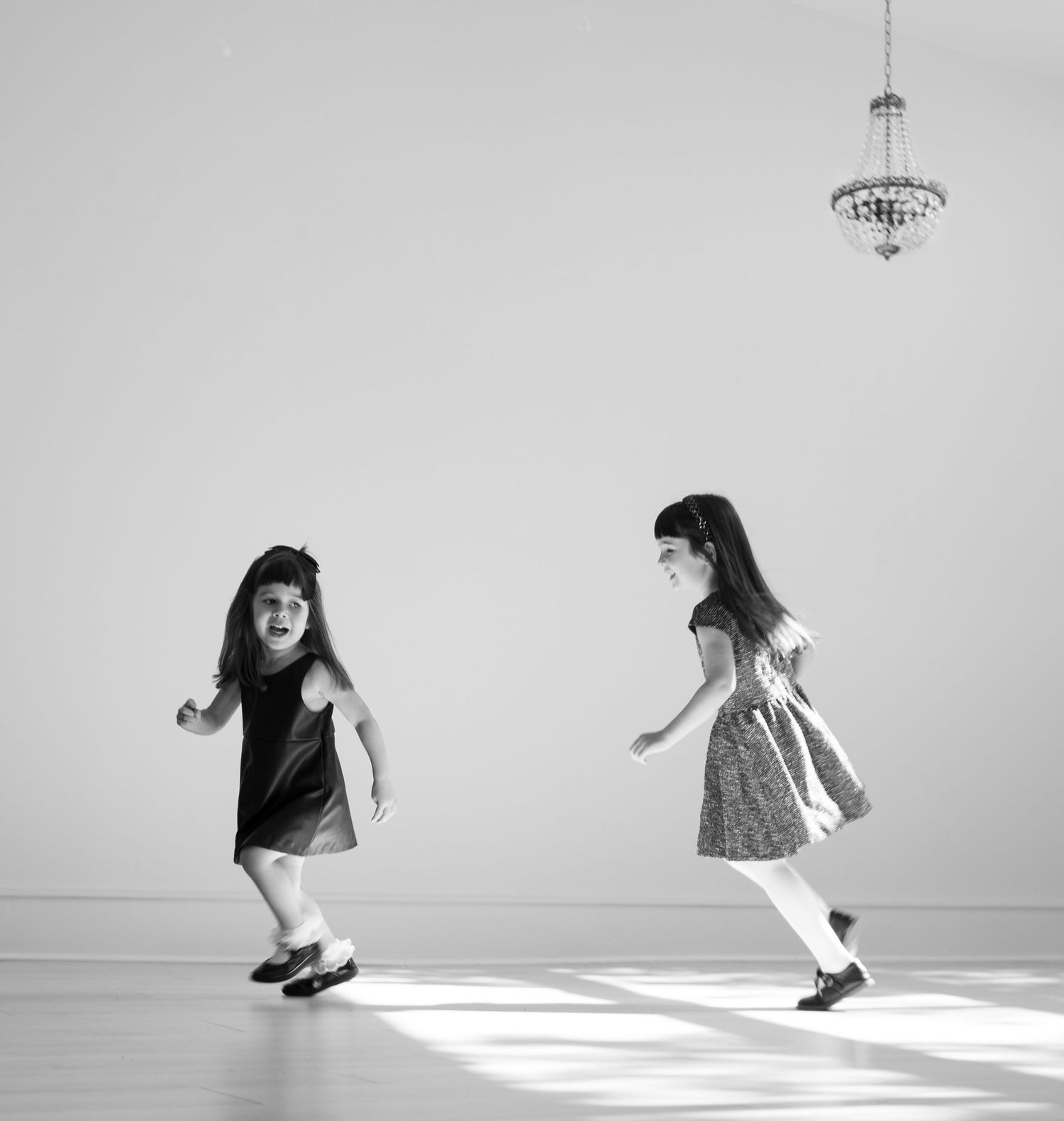 Black and white photo of two little girls dancing joyfully under a chandelier in a white room.