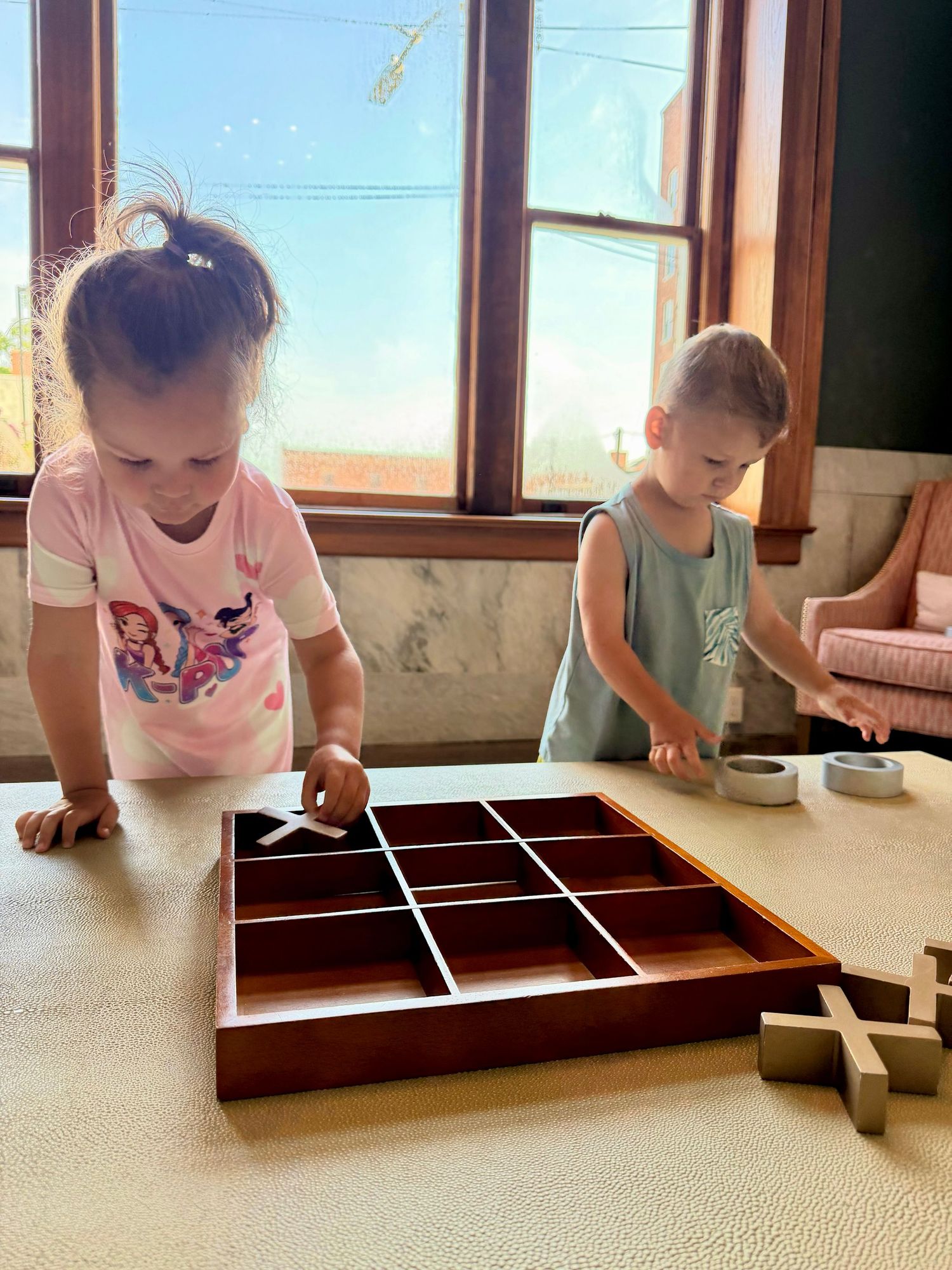 Two young children playing a wooden tic-tac-toe board game on a table near a sunny window.