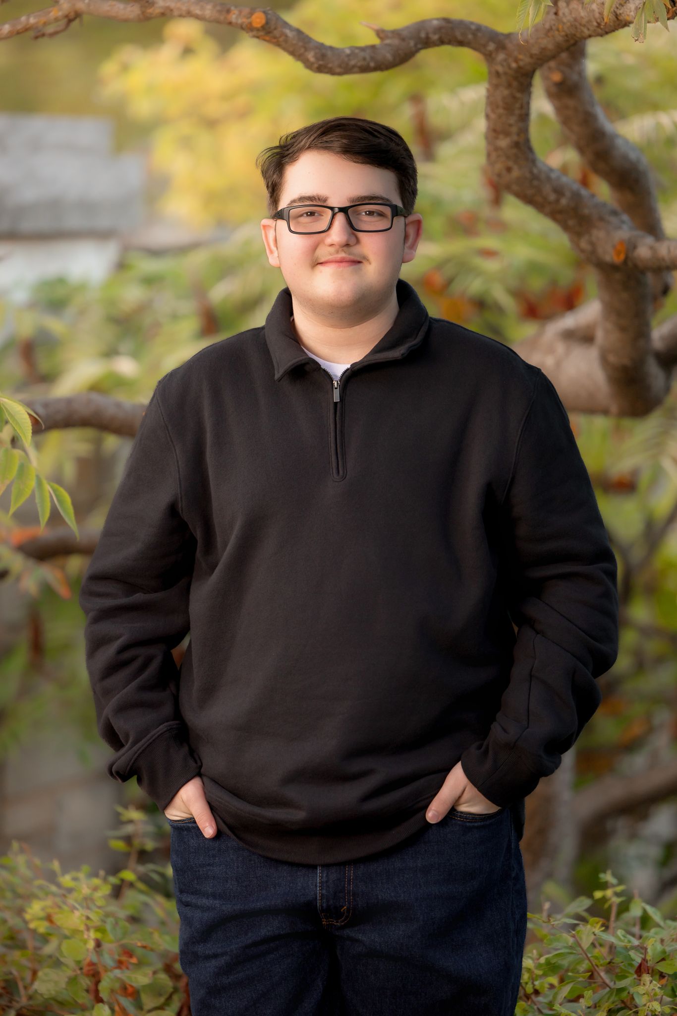 Teen boy high school senior at Frame Park, Waukesha, WI. Golden hour portrait with glasses, warm sunlight, and scenic tree background.