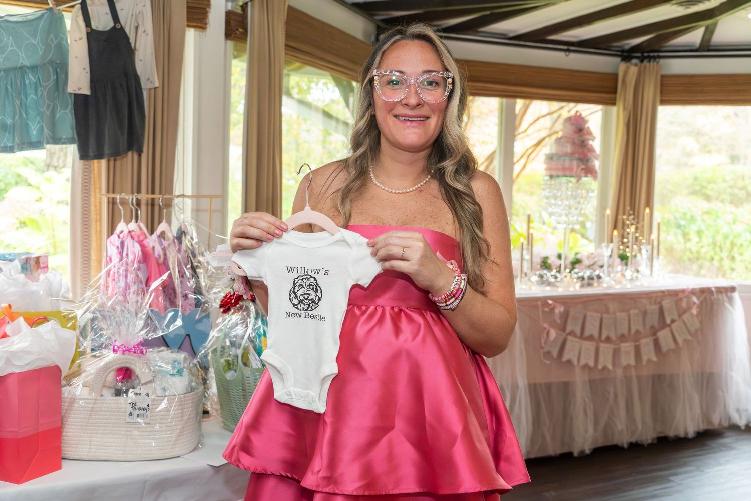 A guest in a pink dress holds up a small white baby onesie at a shower celebration.