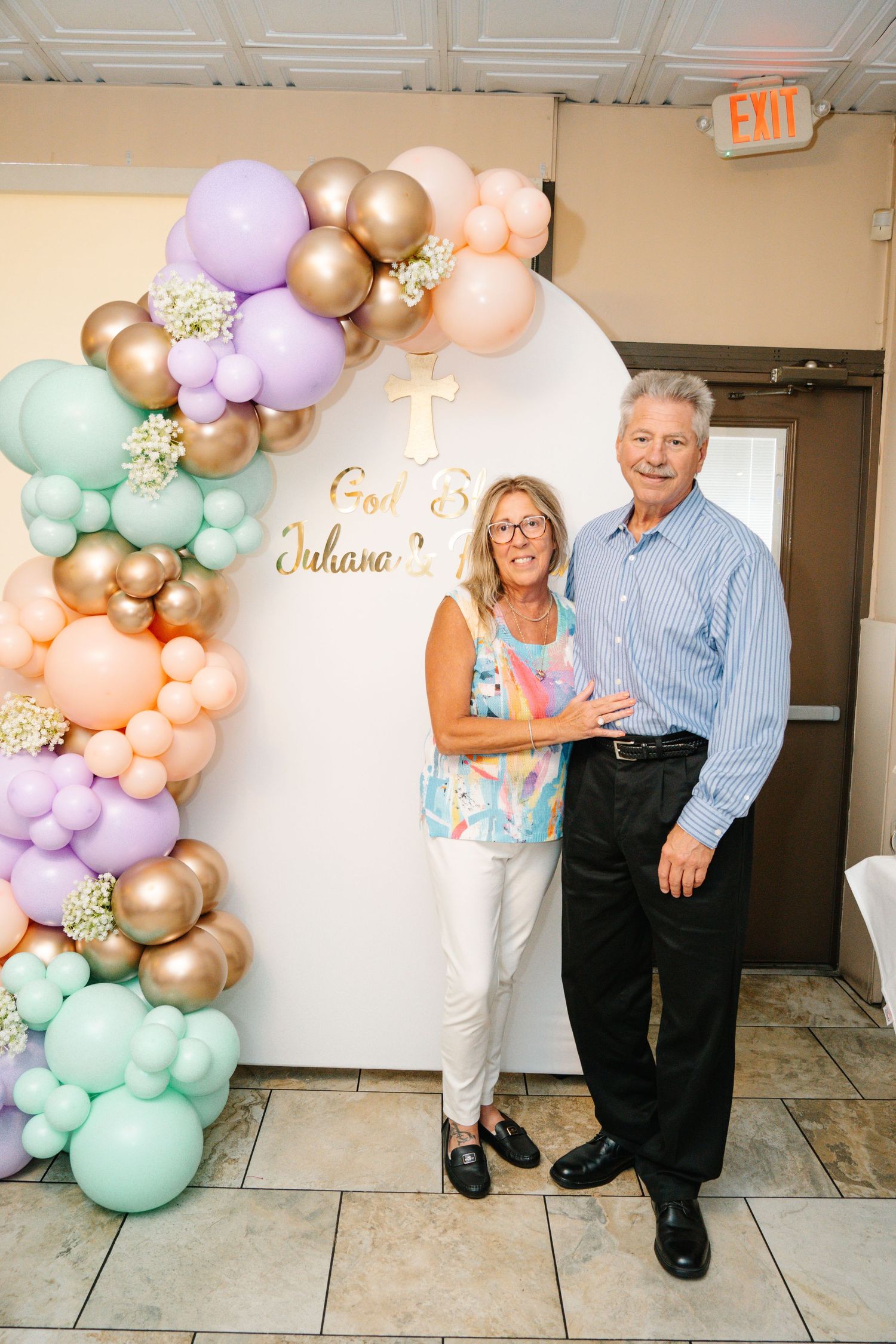 A couple poses by a decorative balloon arch with pastel and gold balloons at a celebration.