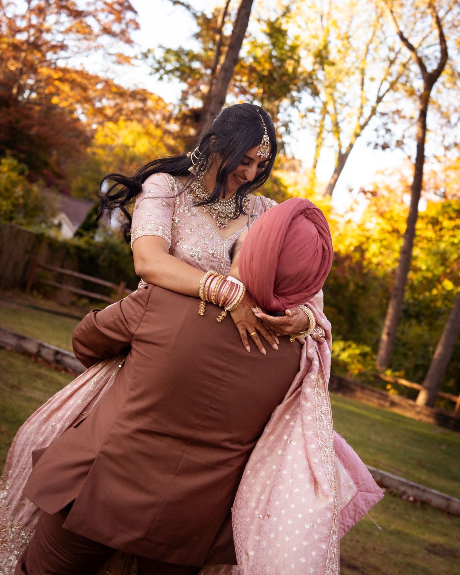 Two people embrace joyfully while wearing traditional South Asian attire in a sunlit autumn setting.