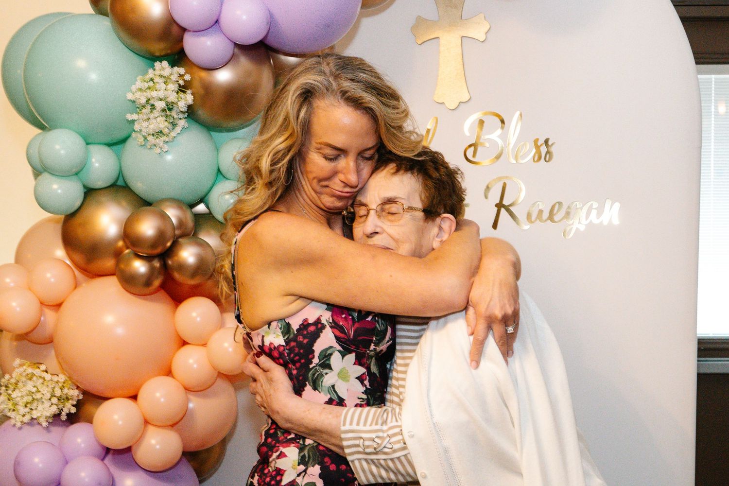 Two people share an emotional embrace in front of a decorative balloon arch at a celebration.