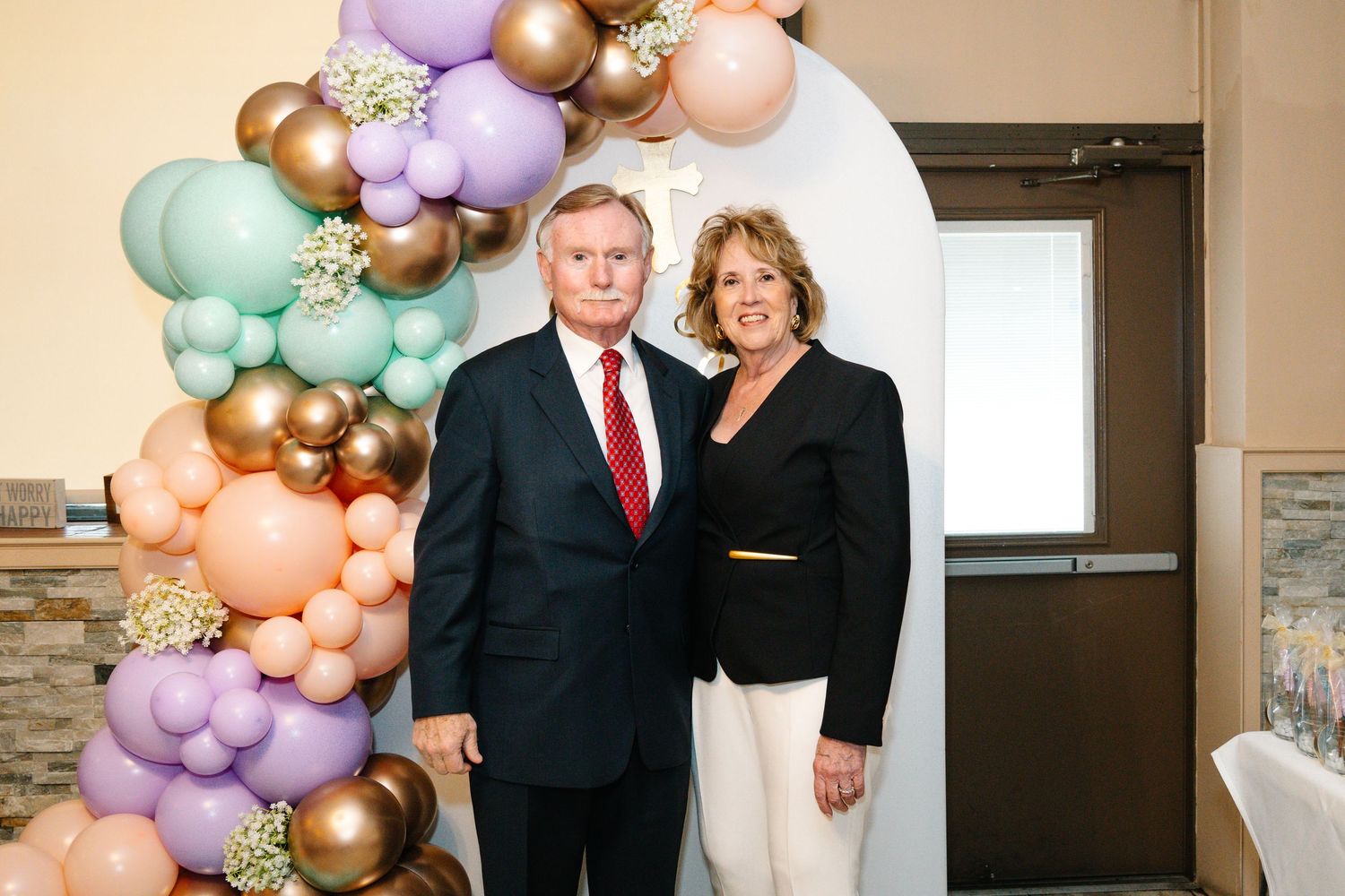 A formal couple stands together in front of a balloon arch at an event.