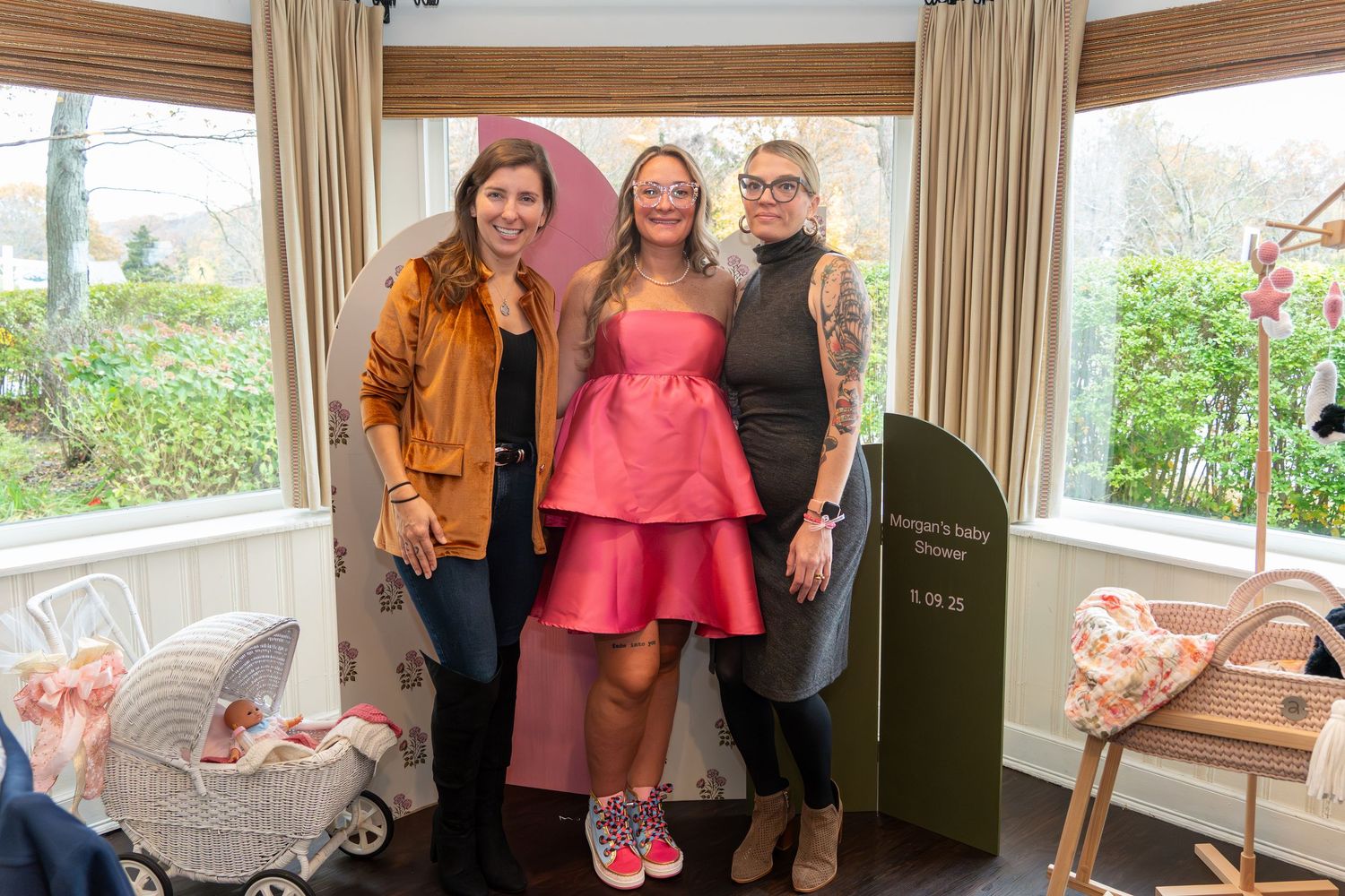 Three guests pose together at a baby shower with pink decor and a baby carriage in the background.