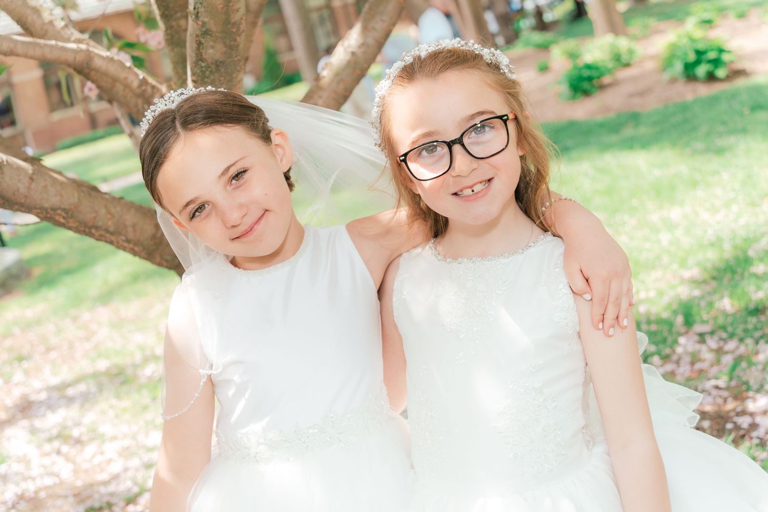 Two people in white dresses pose together under flowering trees.