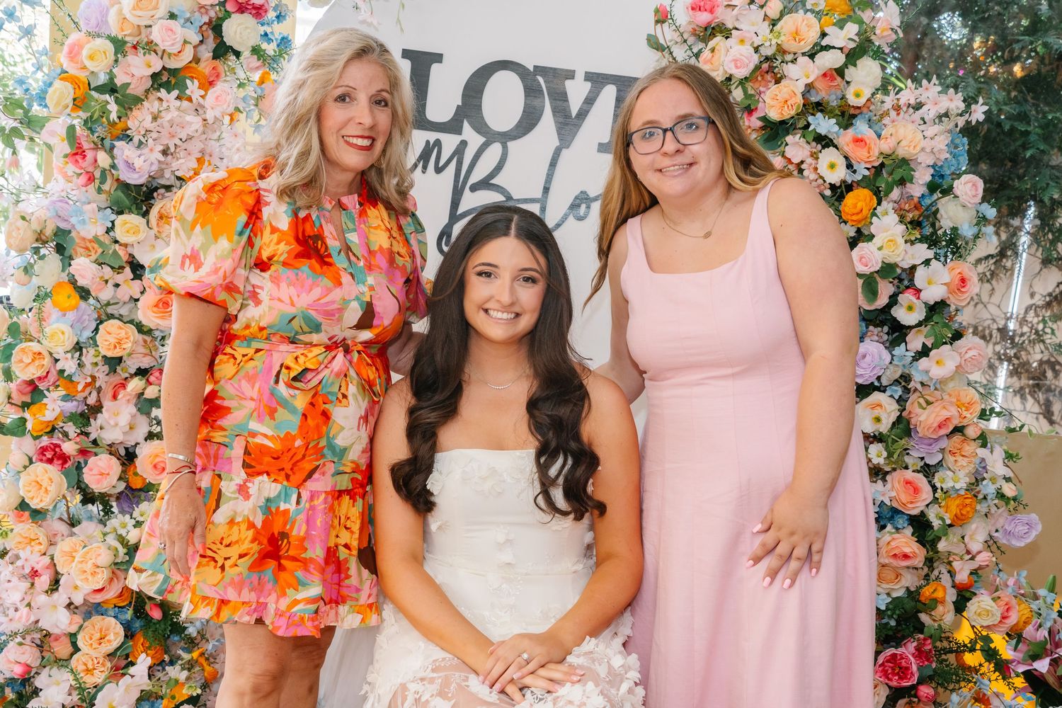 Bridal shower guests pose in front of a floral wall wearing pink and floral dresses.