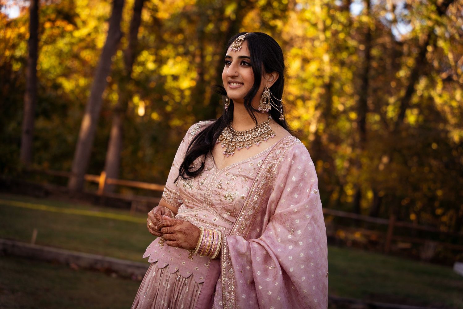 A woman in a pink traditional Indian sari poses elegantly against a backdrop of golden autumn foliage.