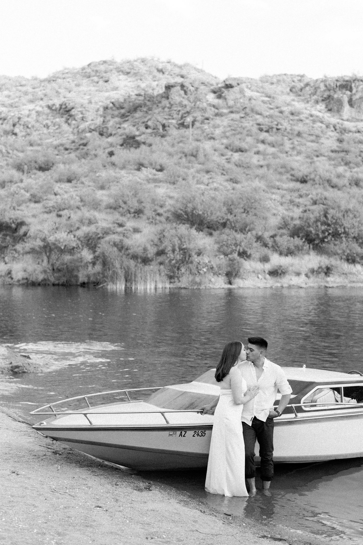 A couple stands near vintage speedboats along a desert lake shore with mountains in the background.