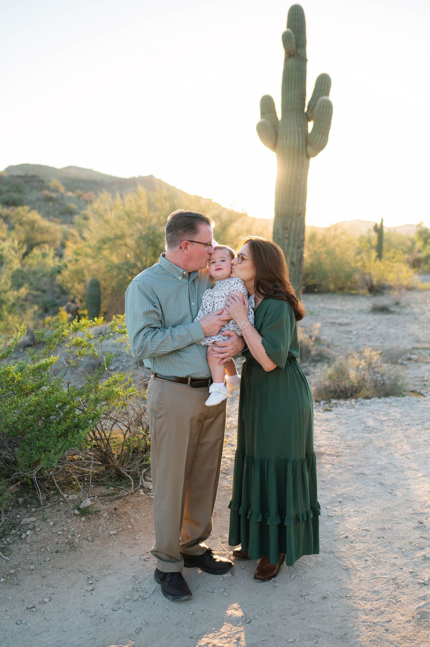 A couple shares an intimate moment in the desert landscape with saguaro cacti and mountains at sunset.