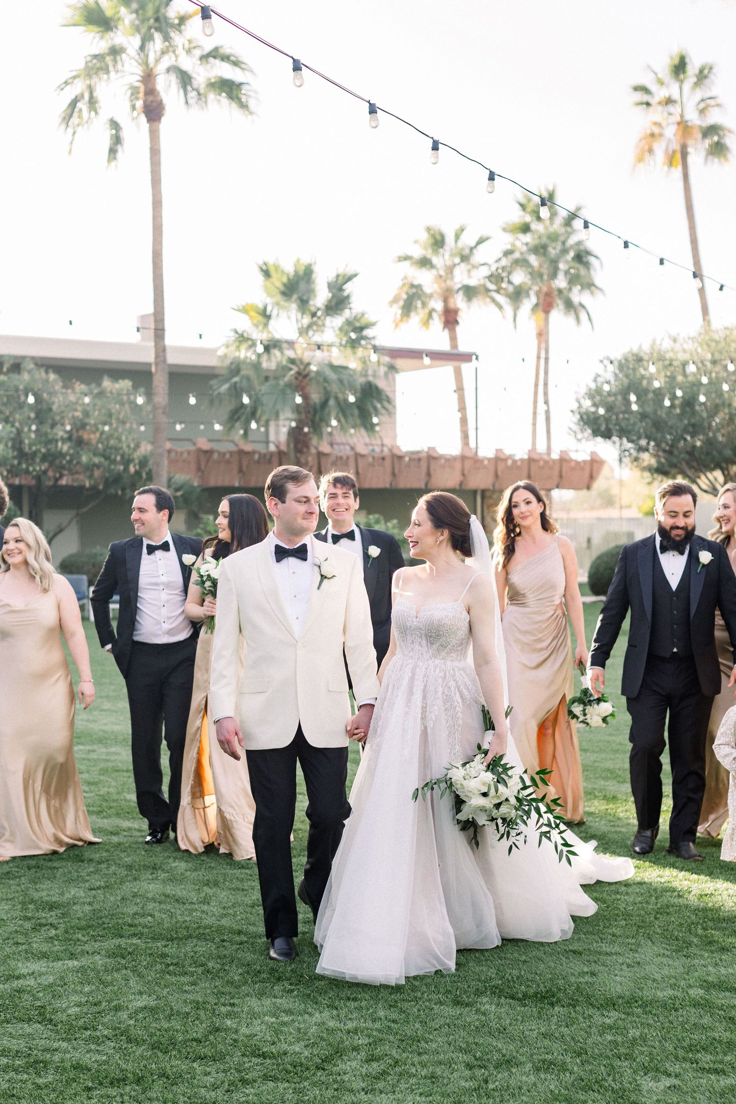 Elegant wedding party walking on lawn with palm trees and string lights overhead.