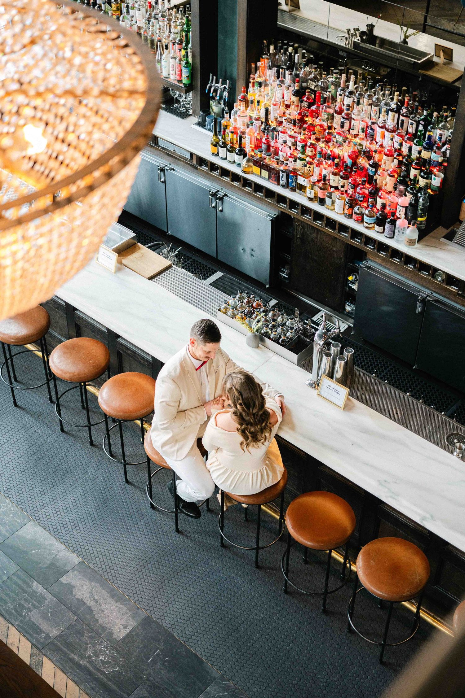 An overhead view of a couple at an elegant bar counter with a stunning chandelier and liquor display.