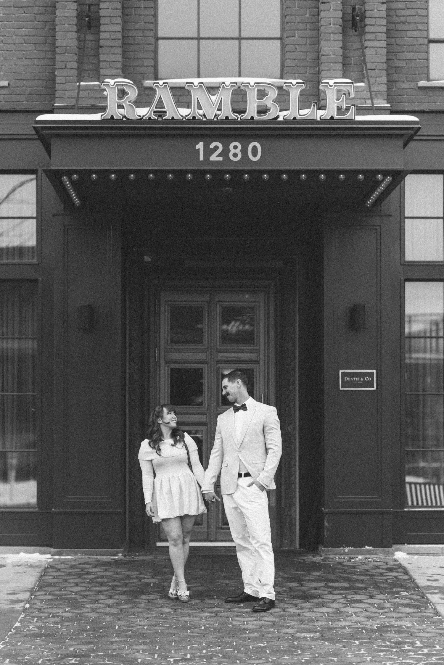 A couple stands beneath the Ramble sign outside a brick building entrance.