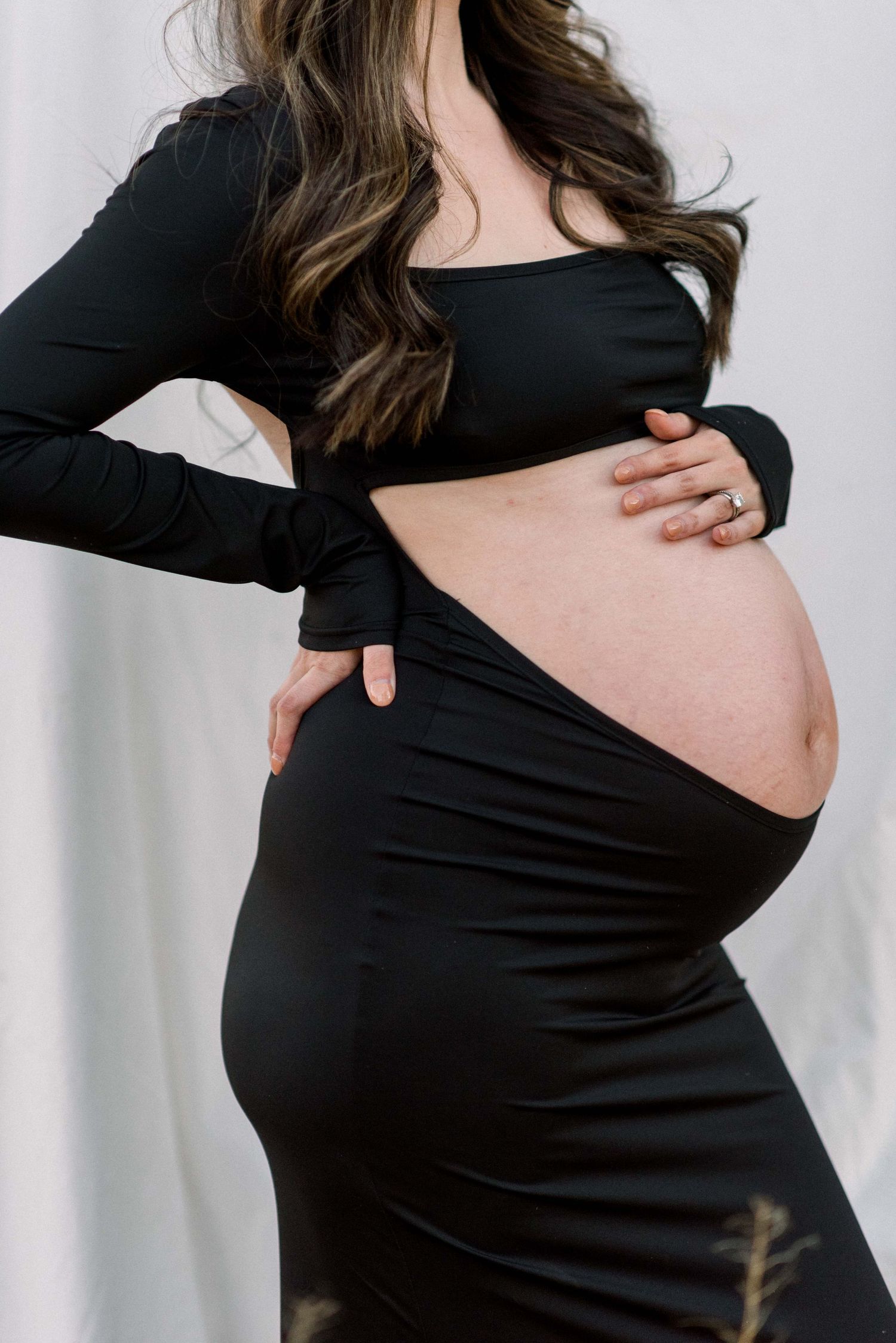 Close-up maternity portrait highlighting baby bump with black form-fitting dress against light background.