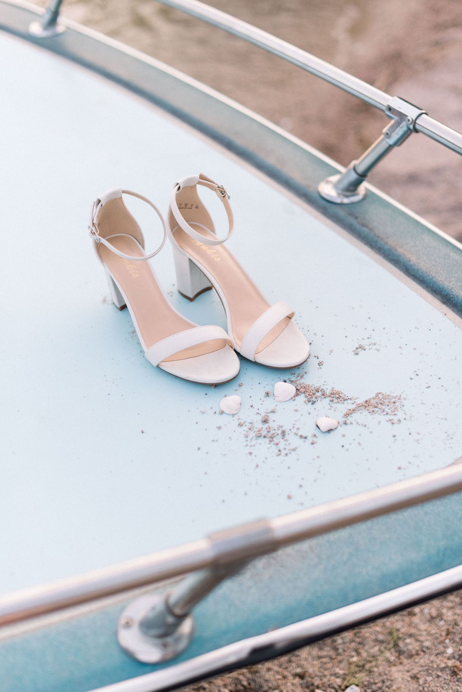 White strappy heeled sandals placed on a light blue surface with metallic railings in the background.