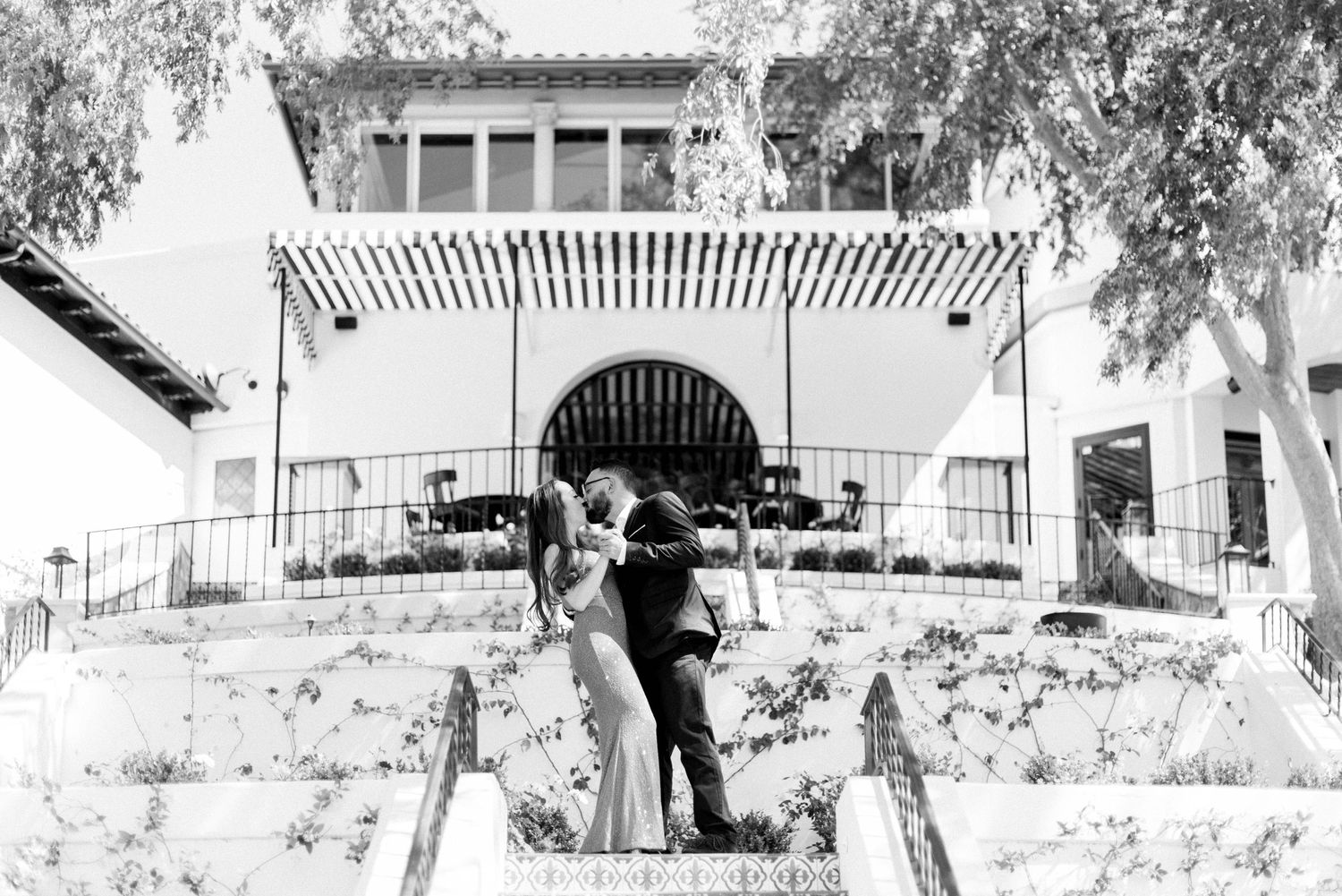 A romantic couple embraces on outdoor steps leading to a Spanish-style building with archways and pergola.