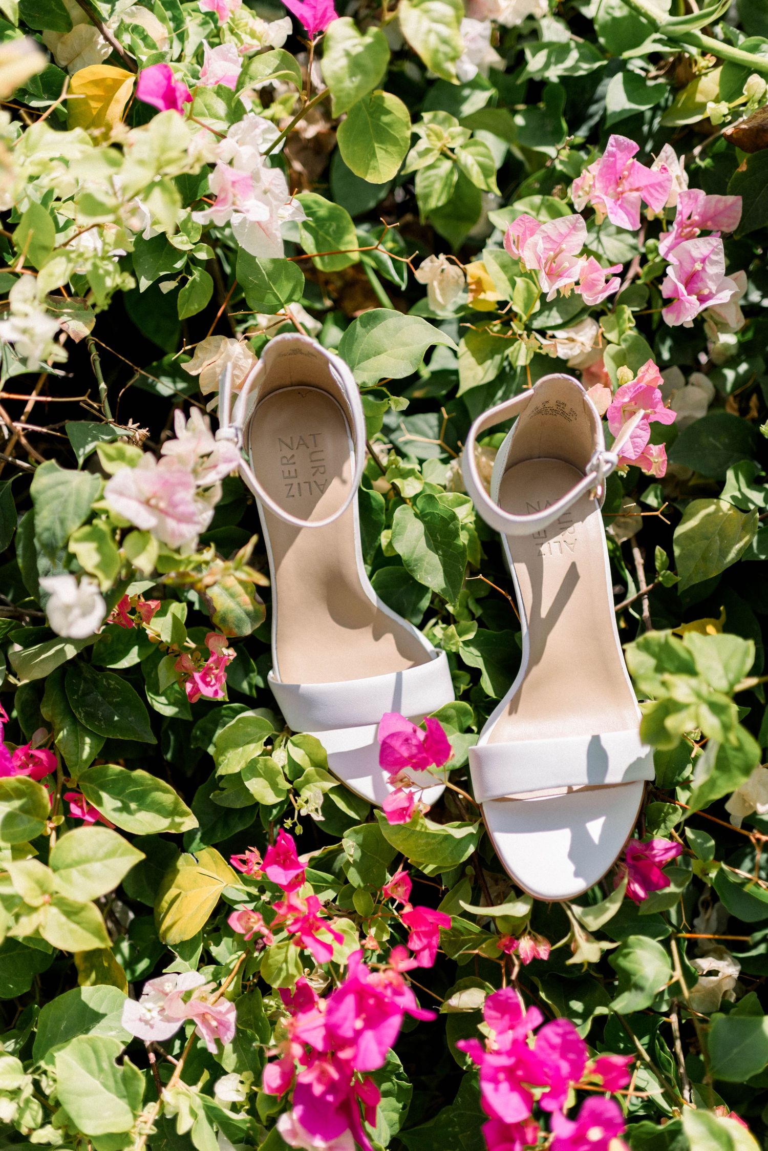 White bridal sandals artistically placed among pink bougainvillea flowers and green foliage.