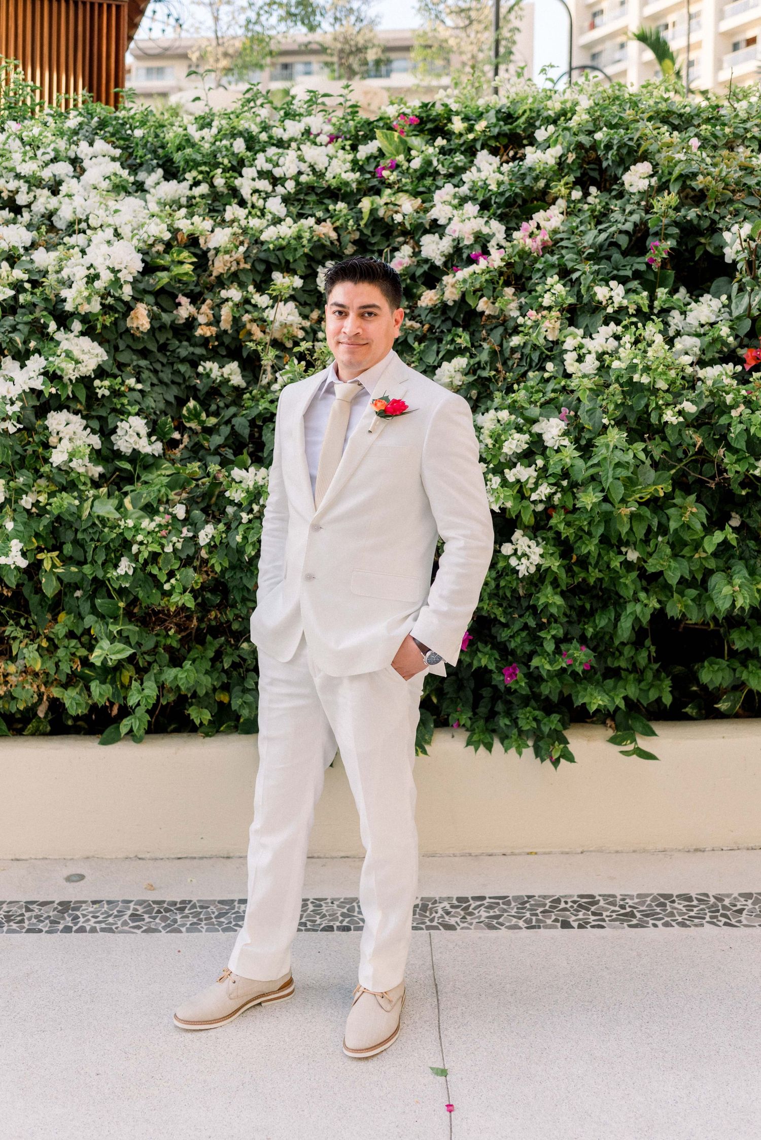 Groom in crisp white suit poses against wall of climbing roses at tropical venue.