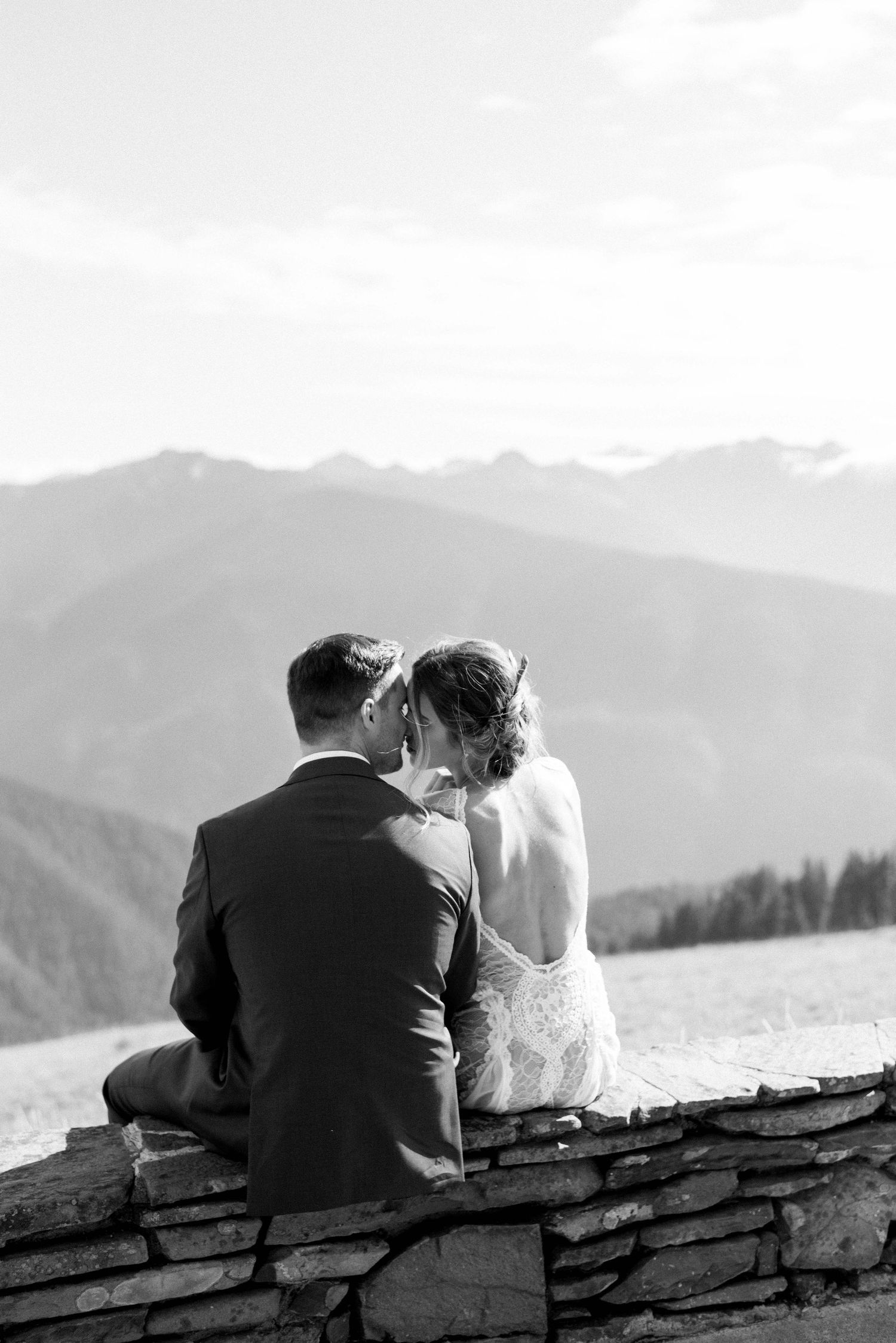 A couple sits intimately on a stone wall overlooking misty mountain ranges in black and white.