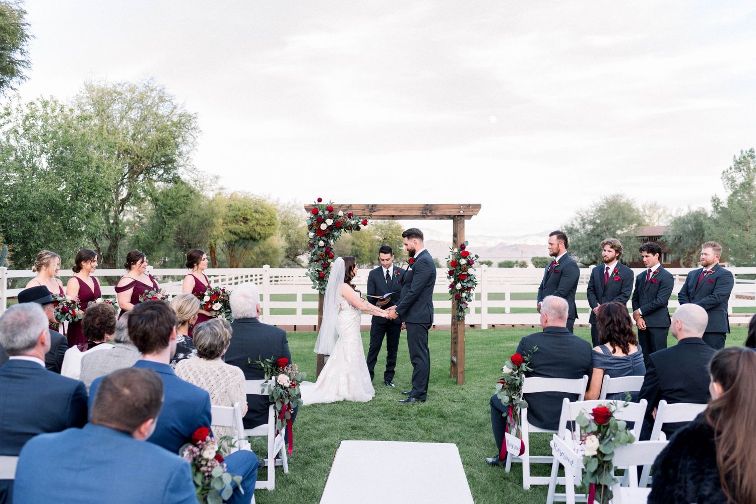 An outdoor wedding ceremony under a wooden arch with white chairs and red floral decorations.