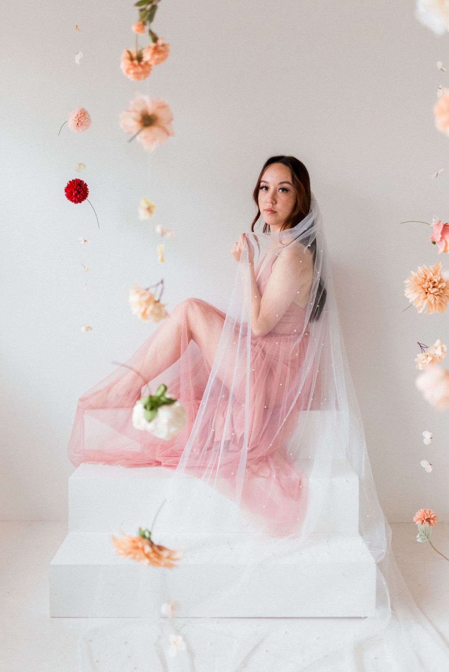 Elegant pose on white platform surrounded by floating peach and pink flowers with flowing veil.