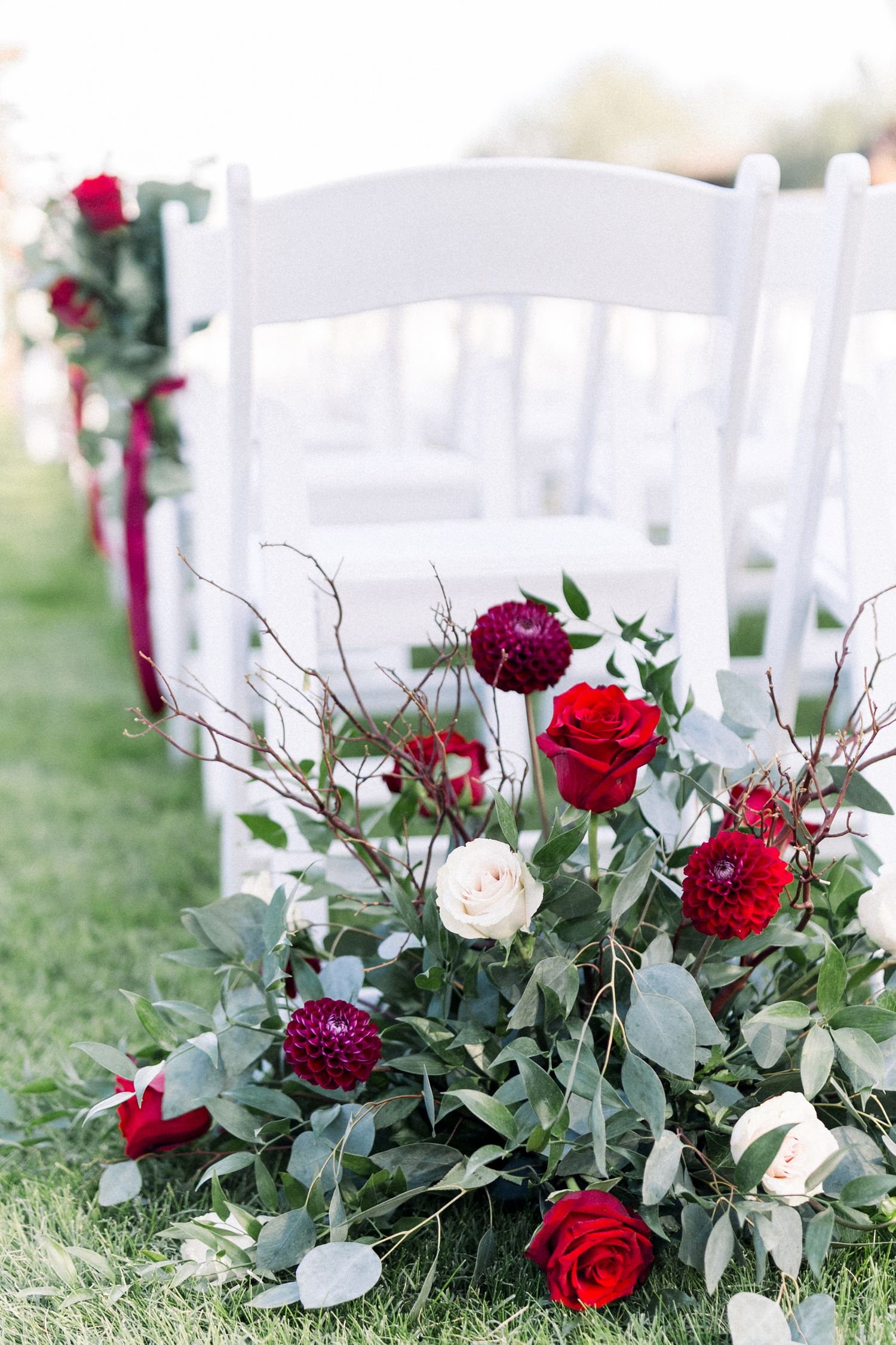 Elegant floral arrangement with red and white roses, eucalyptus leaves beside white wedding chairs.