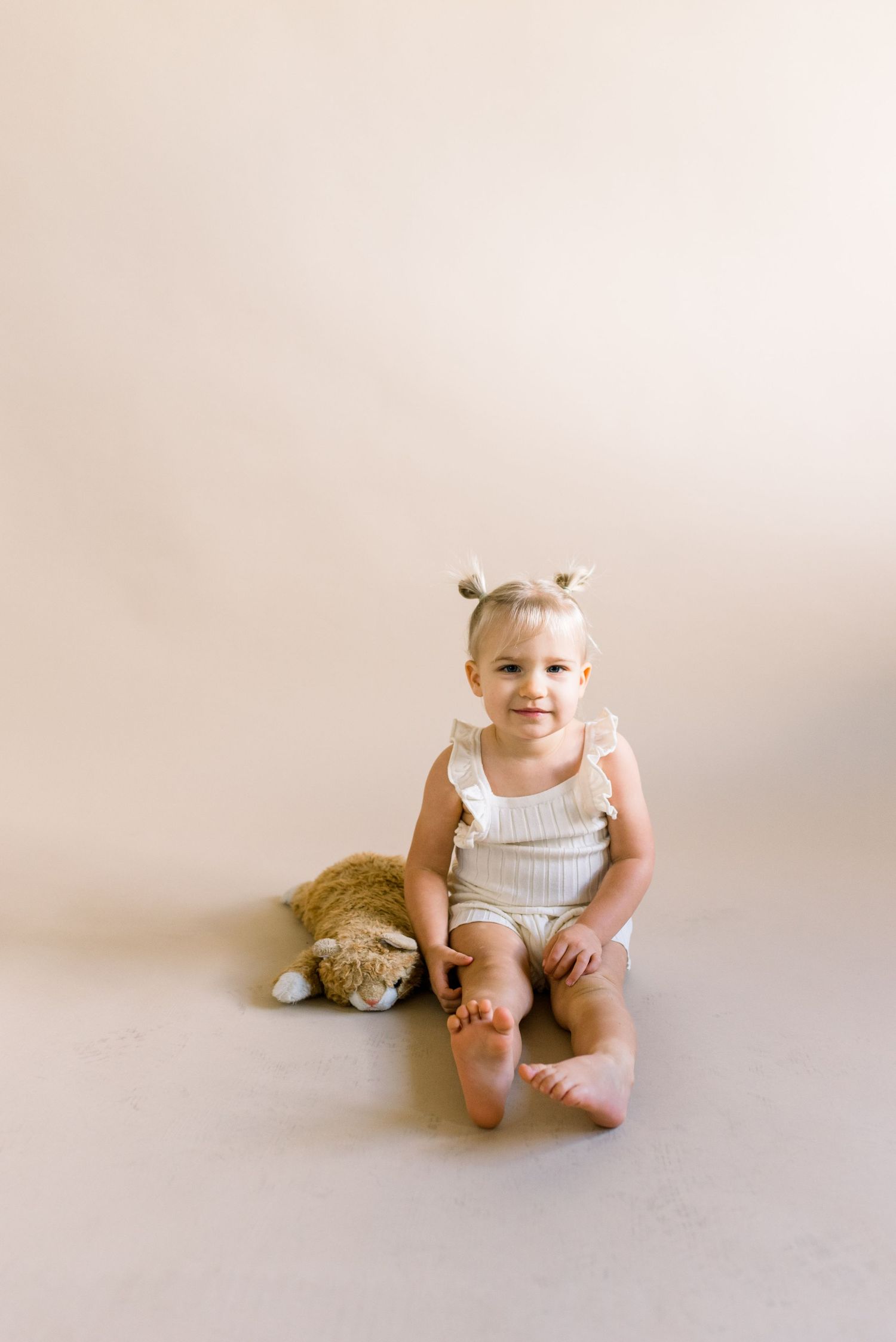 A small child in white outfit sitting with stuffed animal on neutral background.