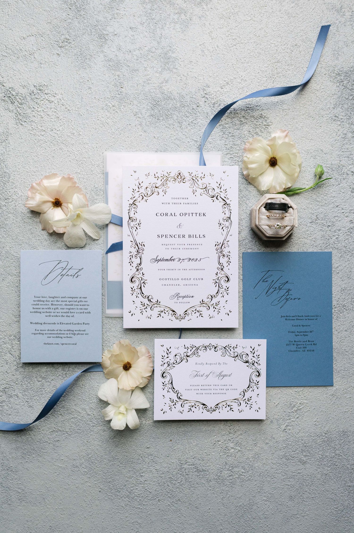 Elegant wedding invitation suite in dusty blue and white with floral details and silk ribbon laid on gray background.