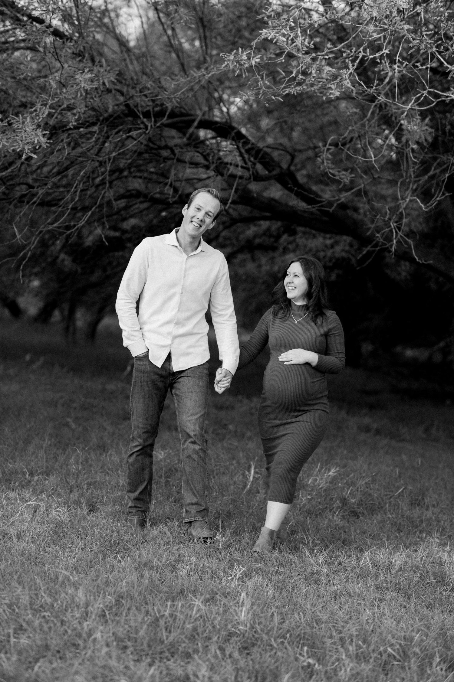 A couple in black and white attire shares playful moments together in a grassy field with trees in the background.