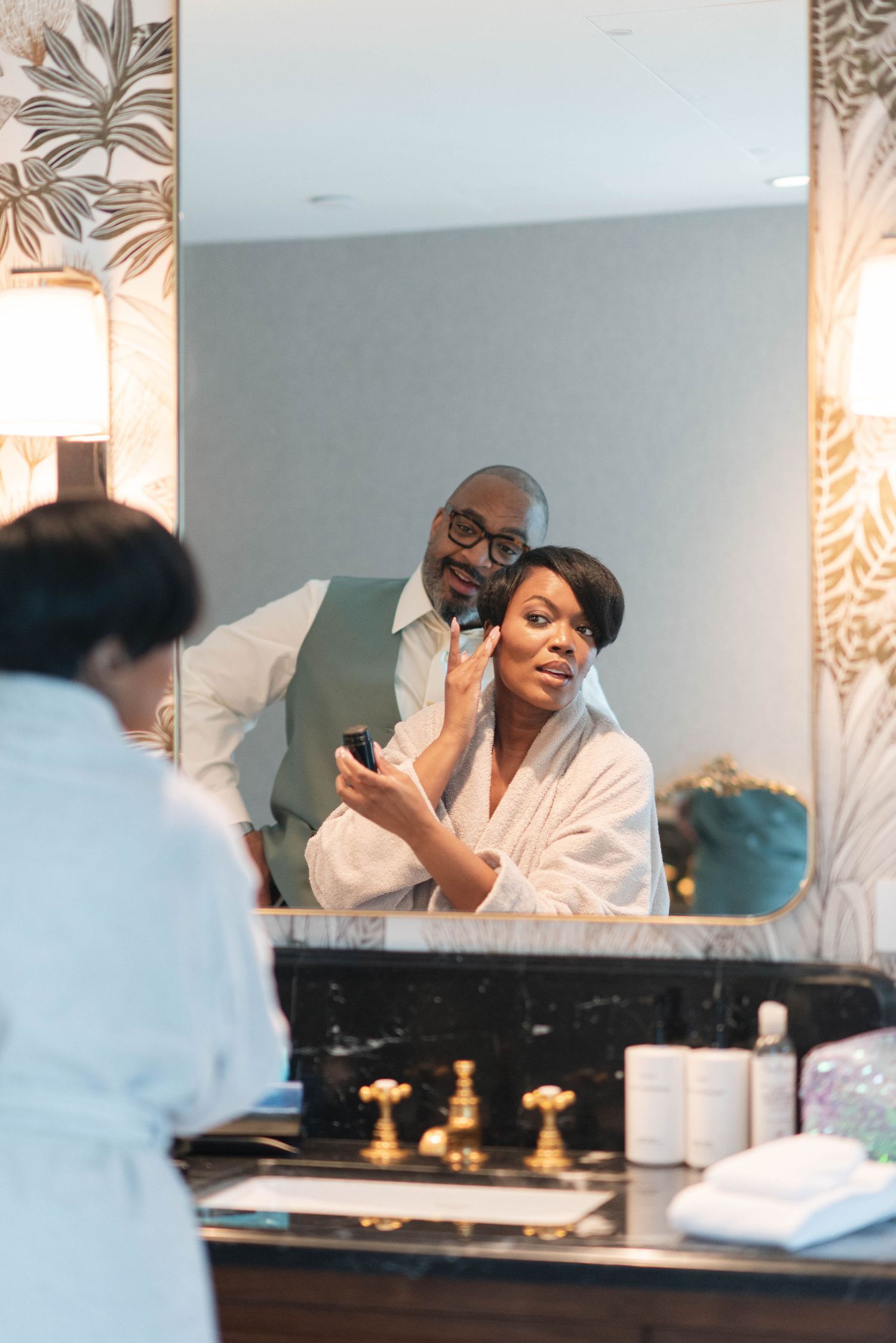 Person in white robe gets ready at bathroom vanity with gold fixtures and palm leaf wallpaper.