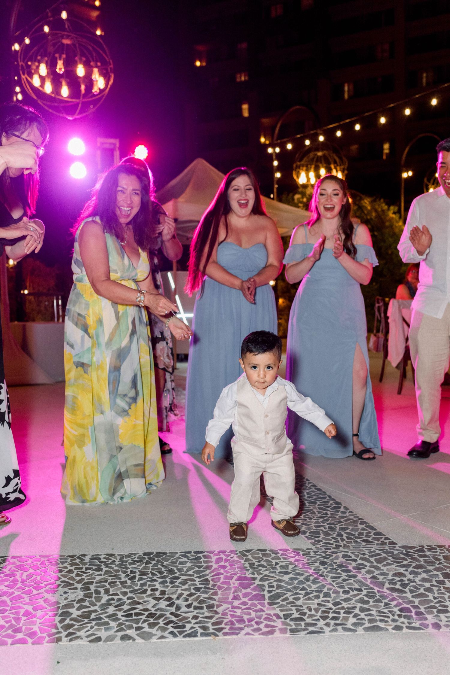 A young boy in white dancing at a wedding reception with colorful lights and guests watching.