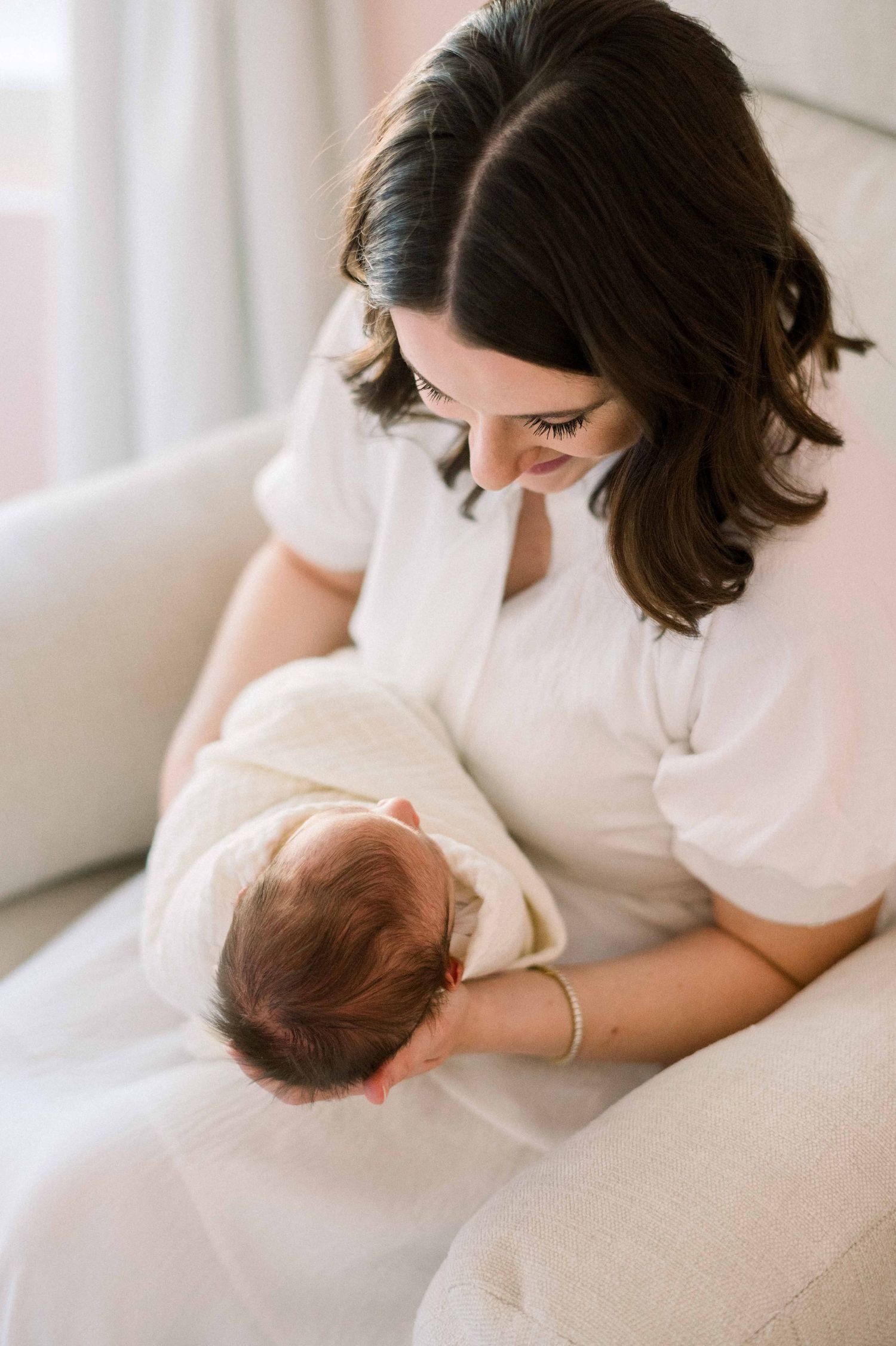 An intimate moment of a woman in white gazing down at her baby on a couch.
