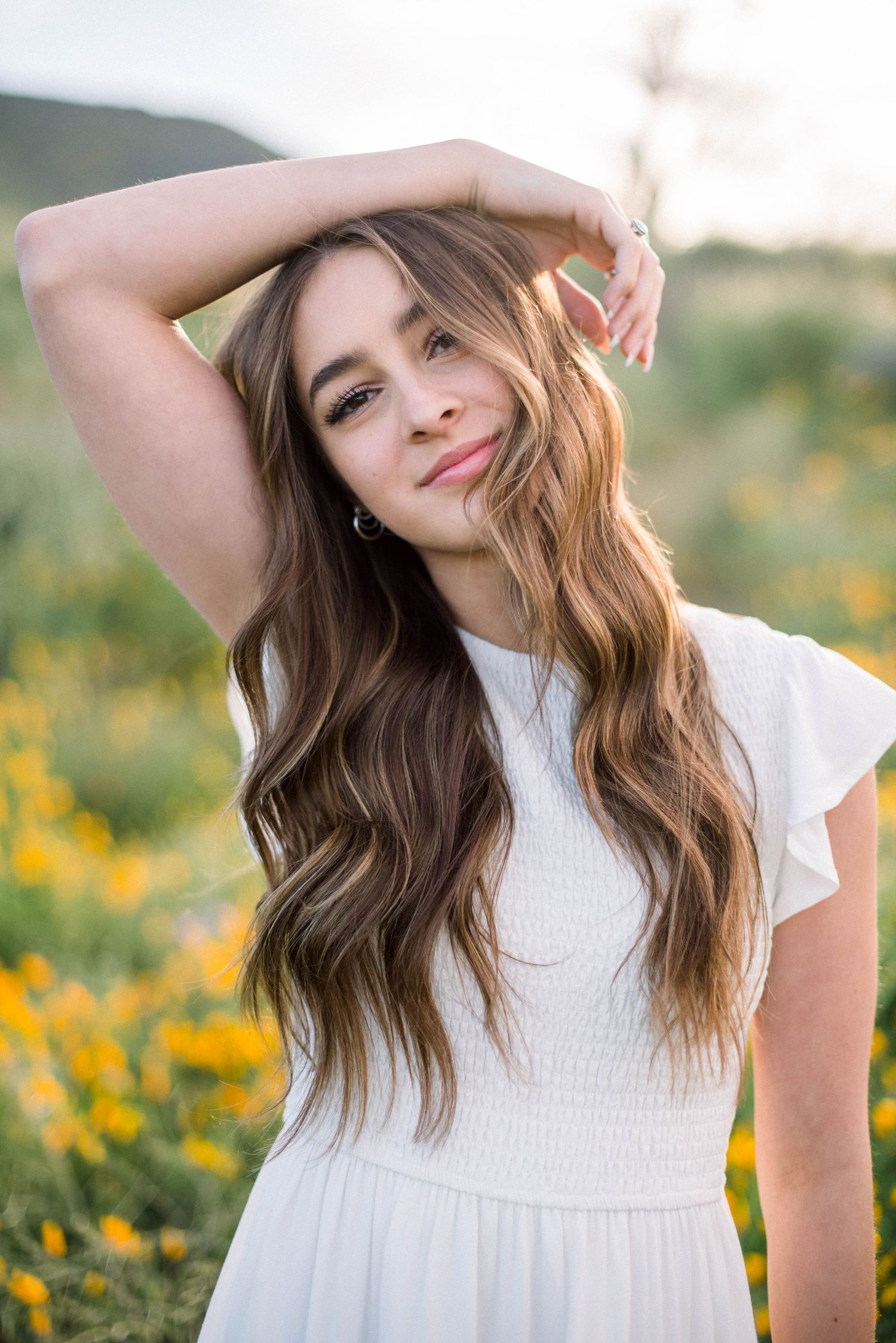 White summer dress with flowing hair against mountain landscape and yellow wildflowers.