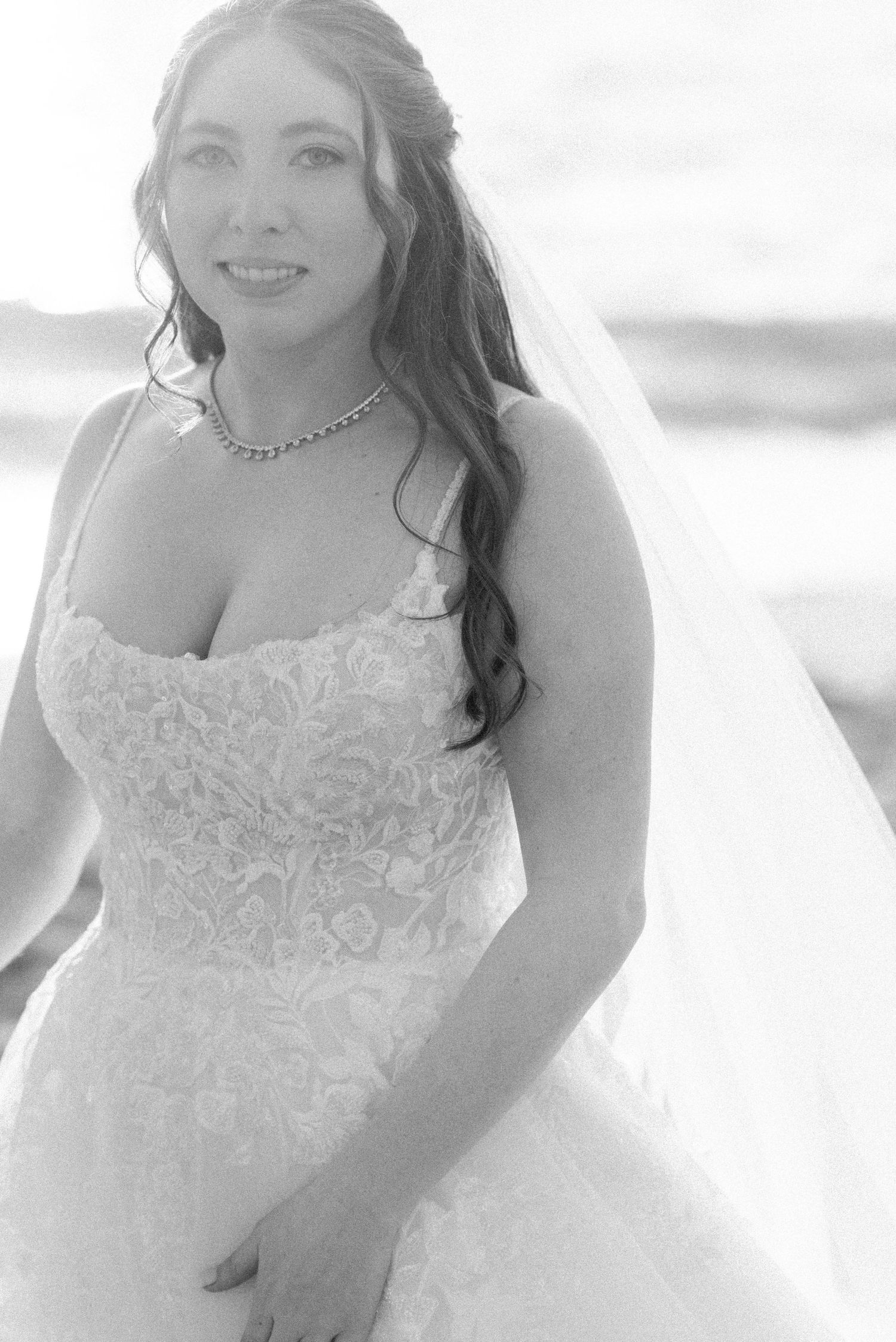 Beautiful bridal portrait in lace wedding gown with veil against ocean backdrop.