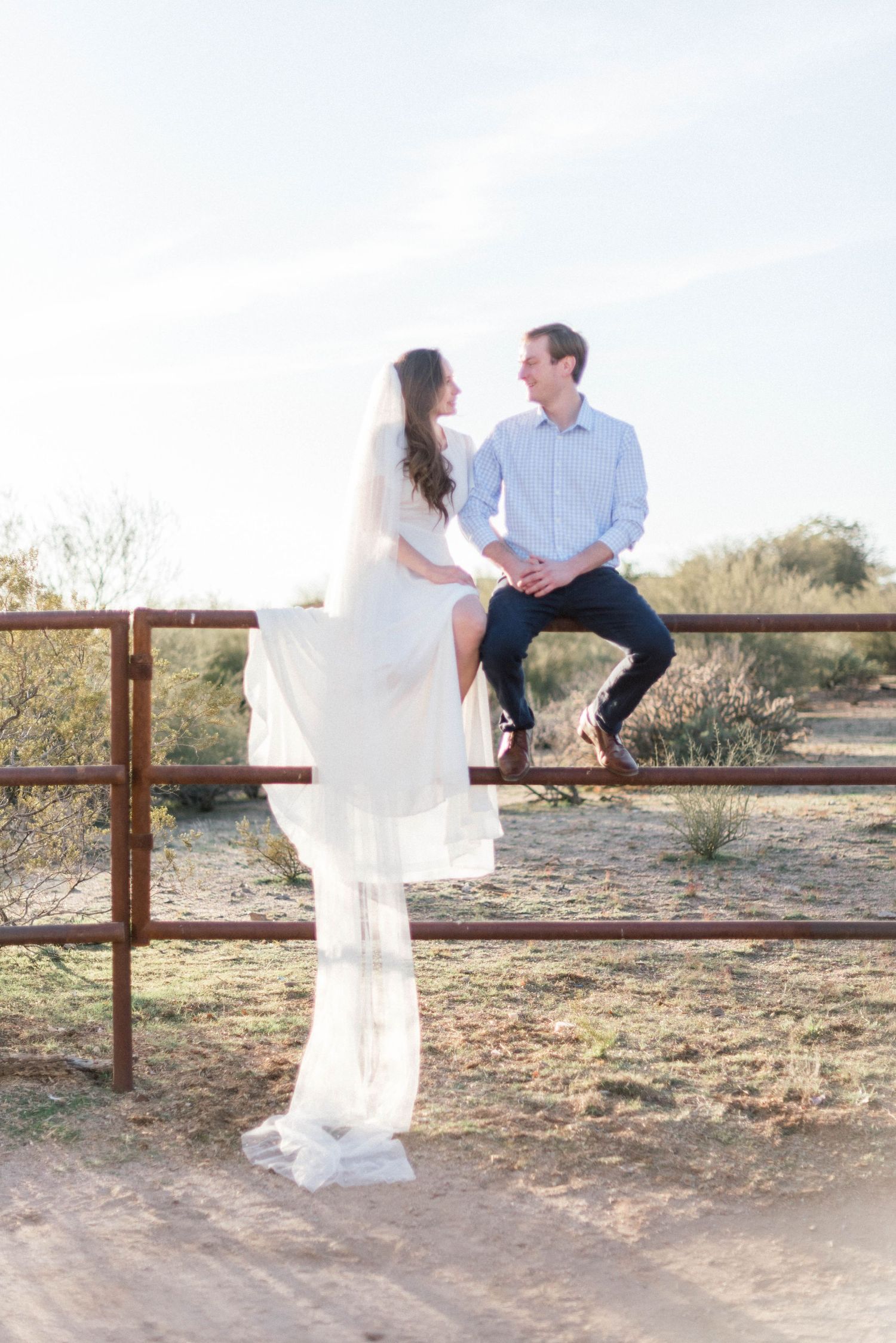 Intimate desert portrait session featuring flowing white dress and casual blue attire on rustic fence.