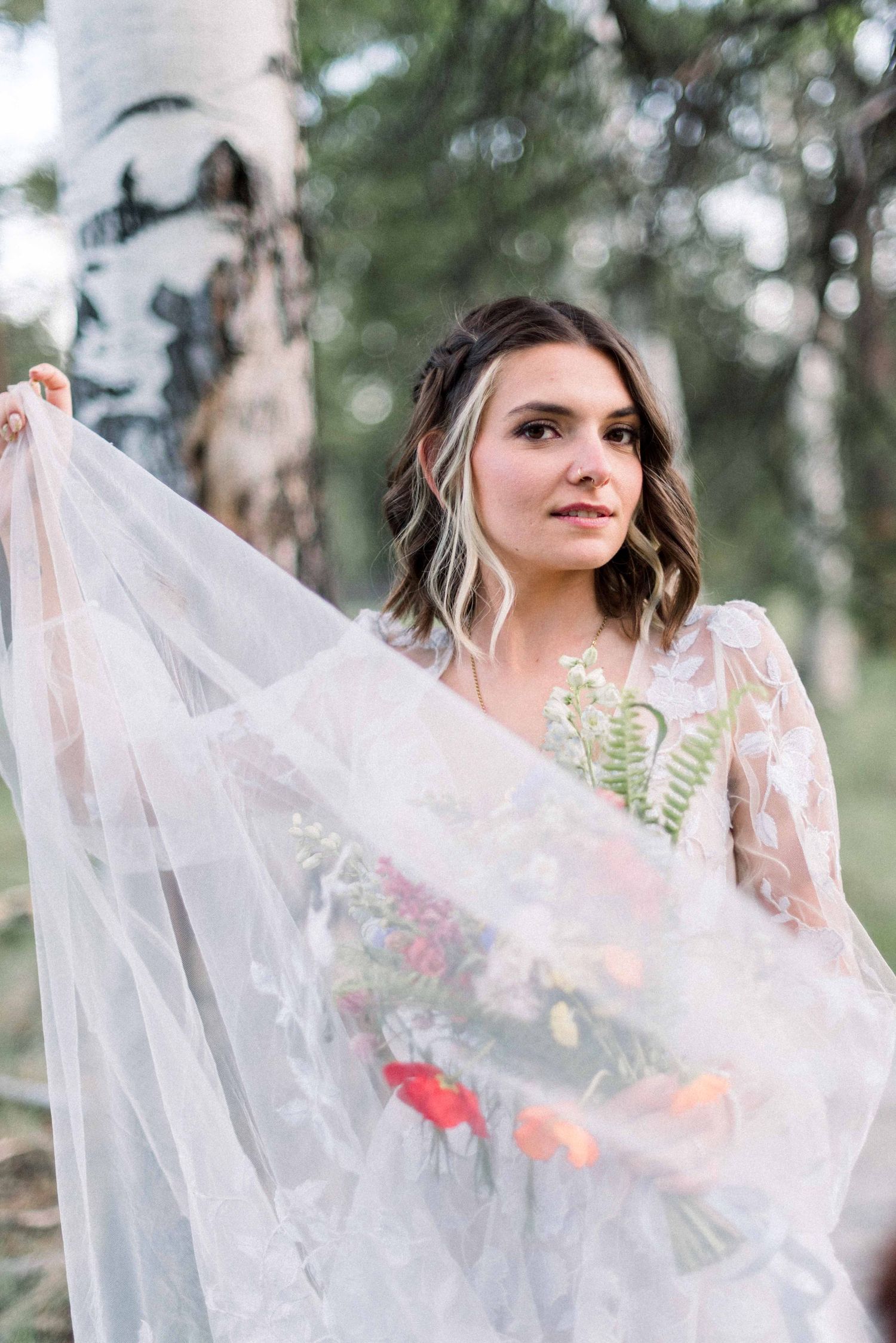 Person in white dress holding sheer veil with floral accents in a forest setting.