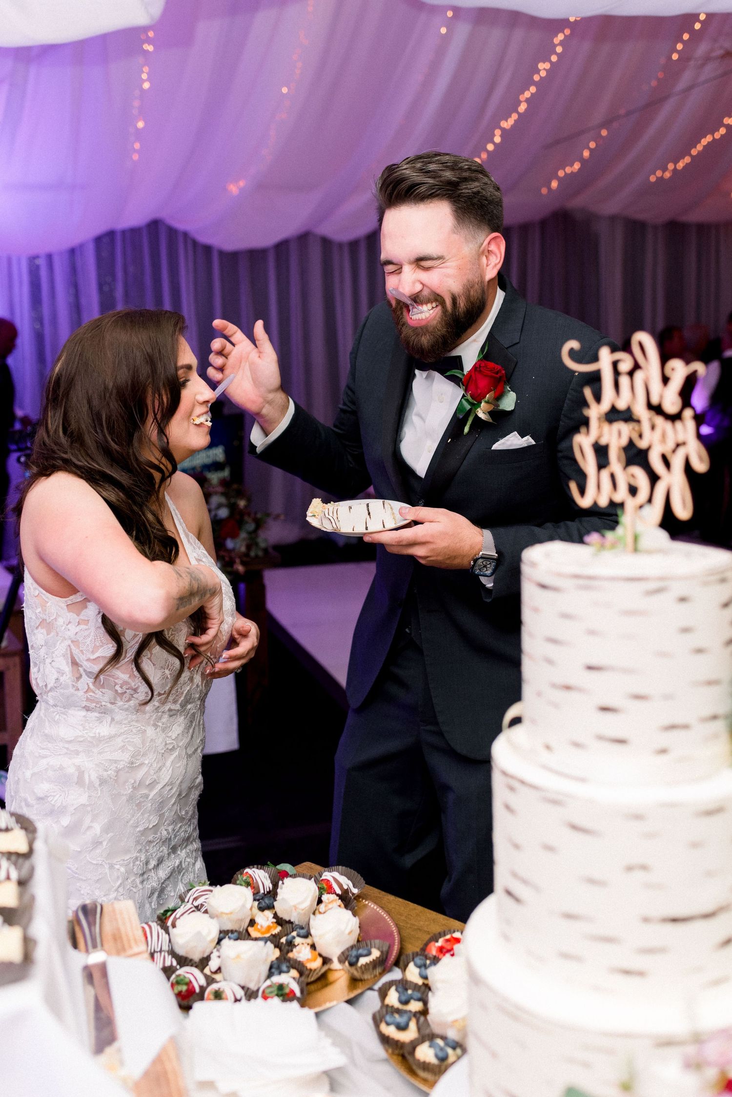 Newlyweds laugh together during cake cutting moment at purple-lit reception.