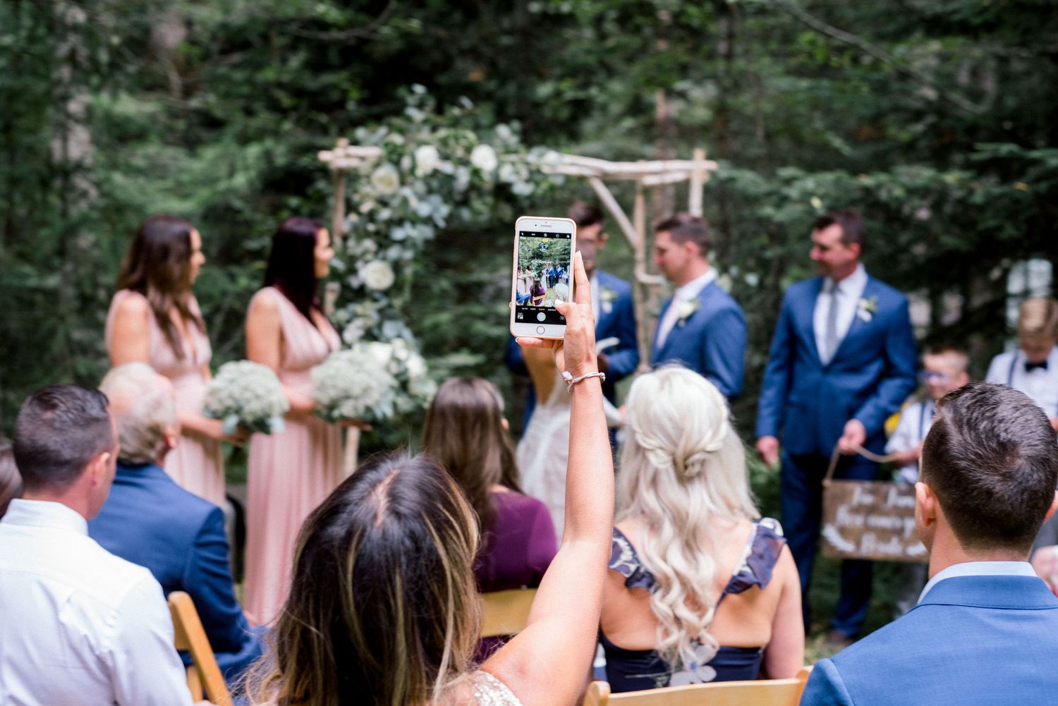 Wedding guests capture ceremony photos on phones during an outdoor forest celebration.