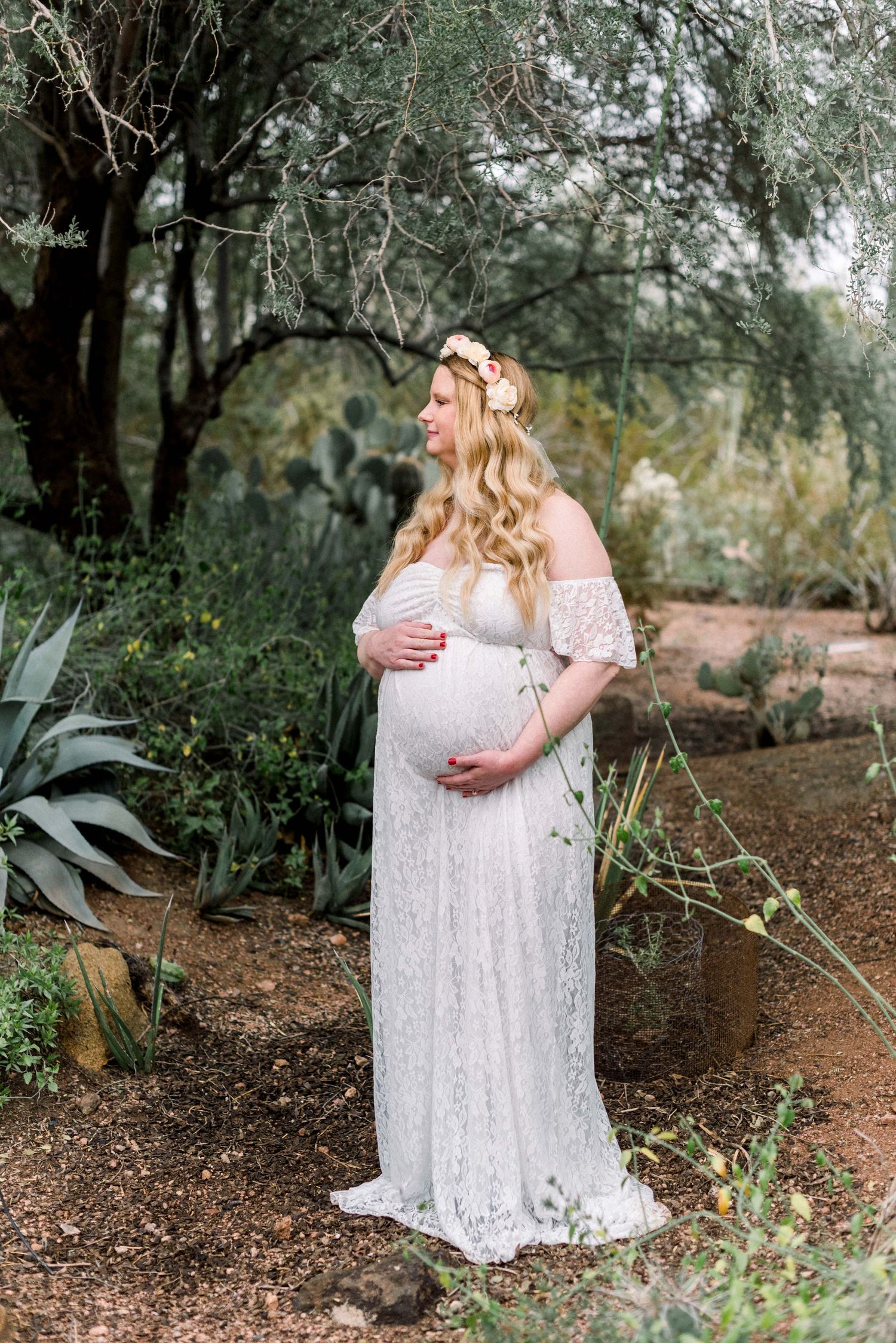 Expectant mother in flowing white dress poses among desert plants and trees.