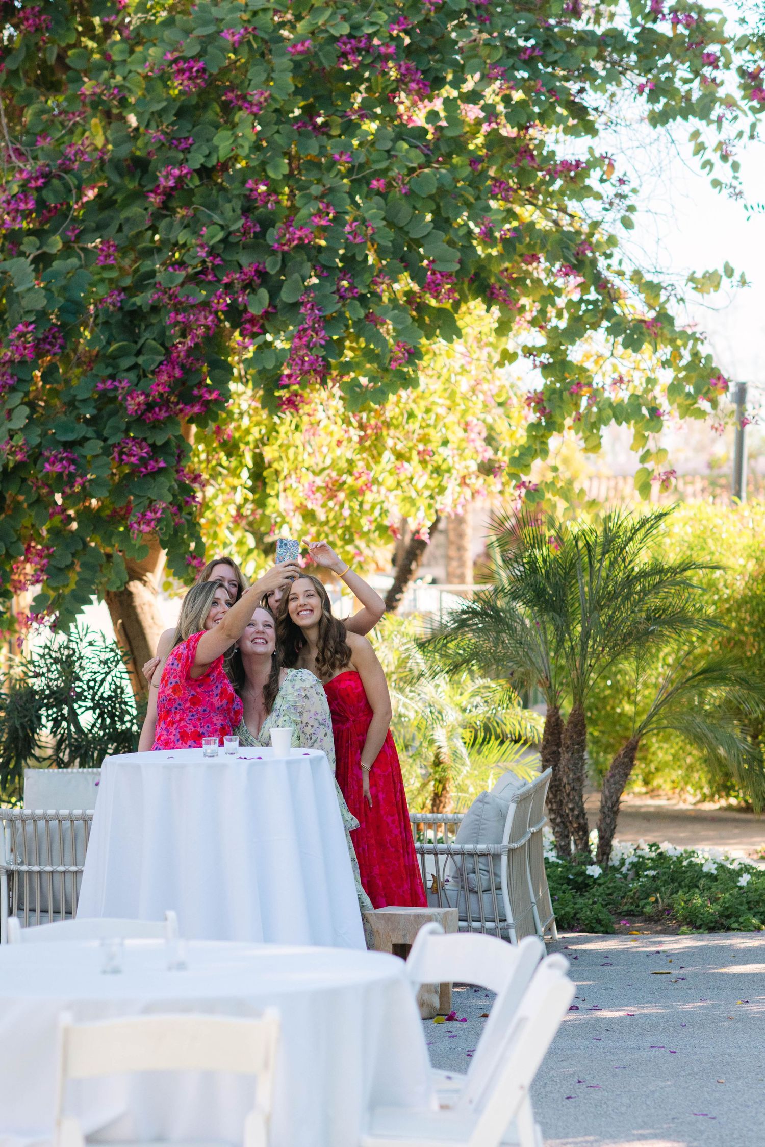 Outdoor wedding reception setup with white tables under flowering trees in a garden setting during golden hour.
