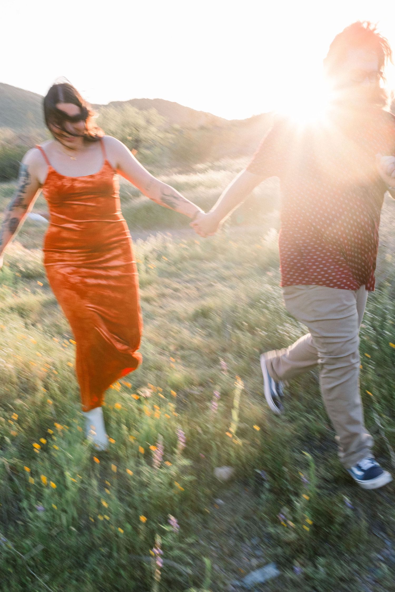 Two people walk through a sunlit meadow with wildflowers while wearing orange and white outfits.